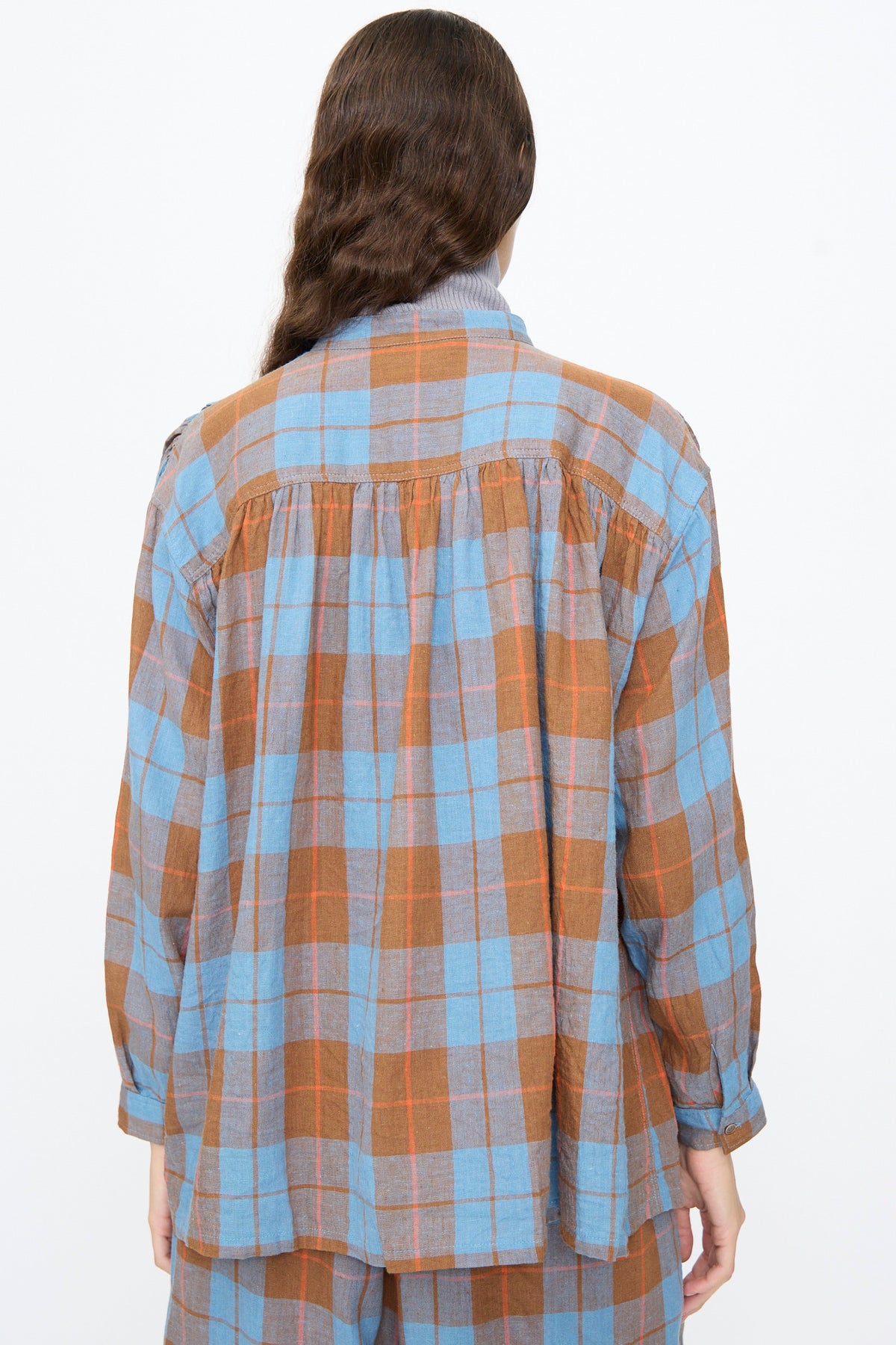 A person wearing an Ichi Antiquités Linen Cotton Check Shirt in Blue, featuring a long-sleeved, band collar design with a blue and brown plaid pattern and a gathered back yoke.