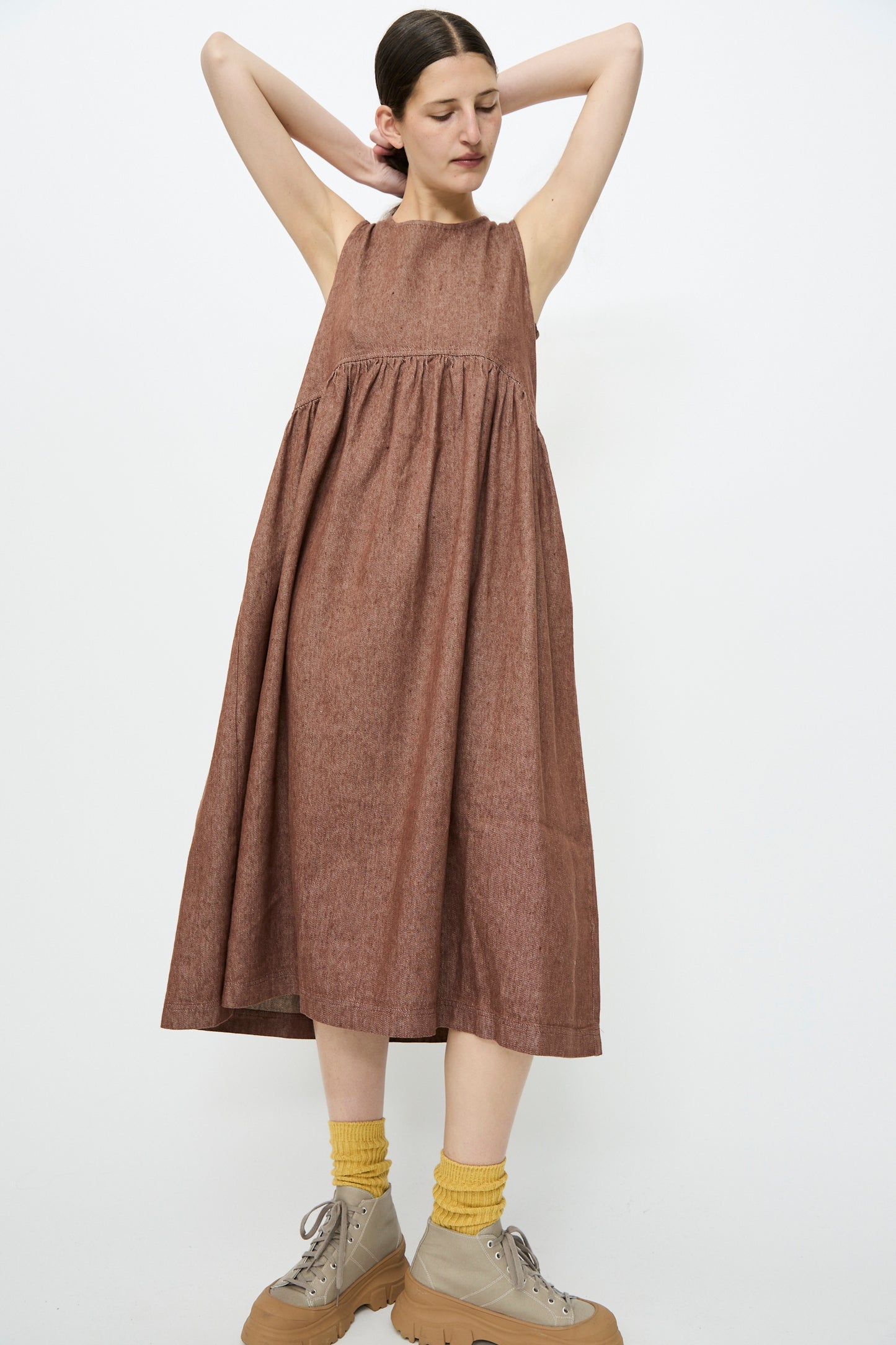 A person in an Ichi Antiquités Linen Cotton Denim Dress in brown, paired with yellow socks and beige platform shoes, stands with arms raised against a white background.
