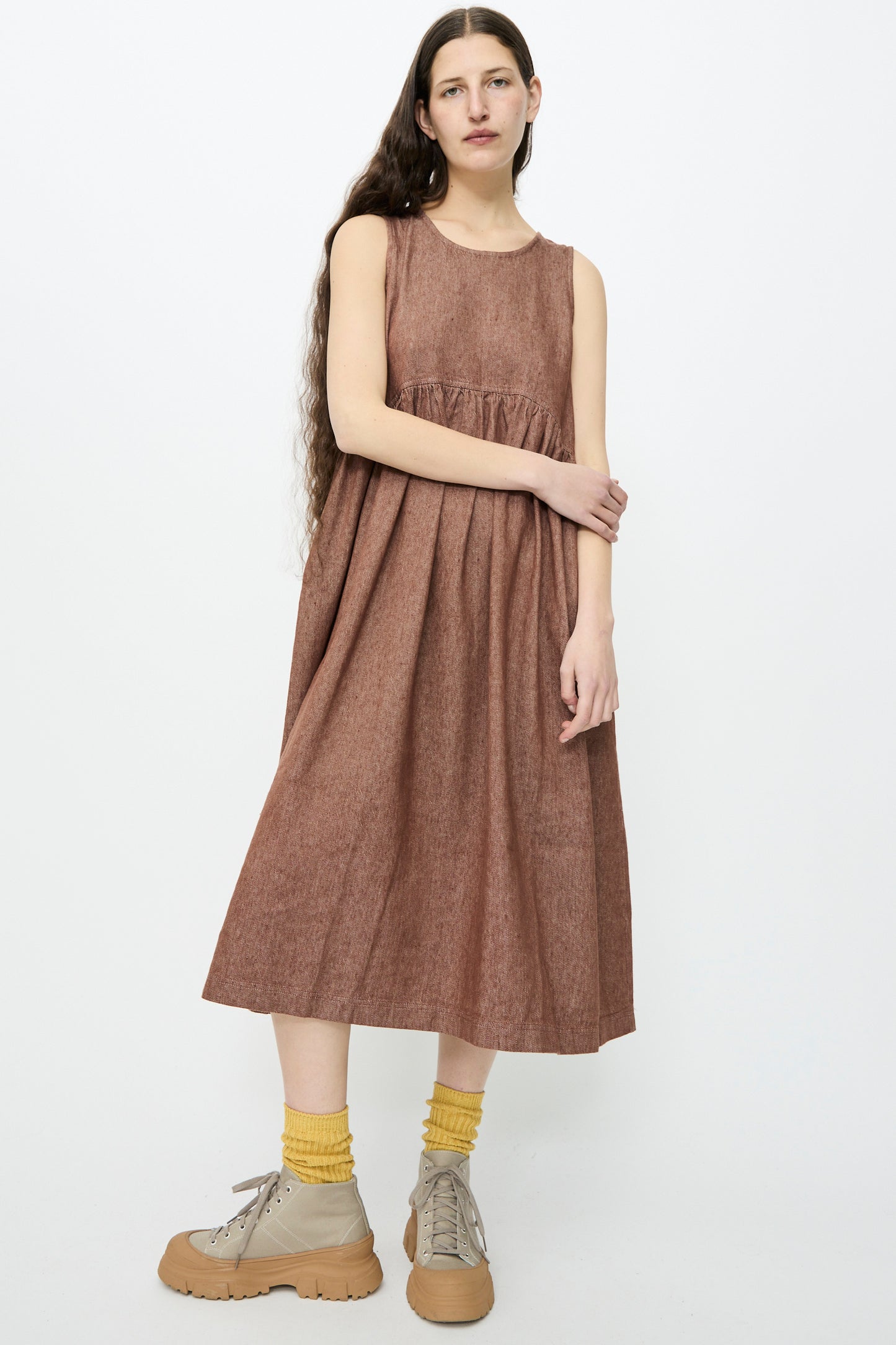 A person with long hair is gracefully standing against a plain white background, wearing an Ichi Antiquités sleeveless relaxed-fit Linen Cotton Denim Dress in brown, paired with yellow socks and beige shoes.