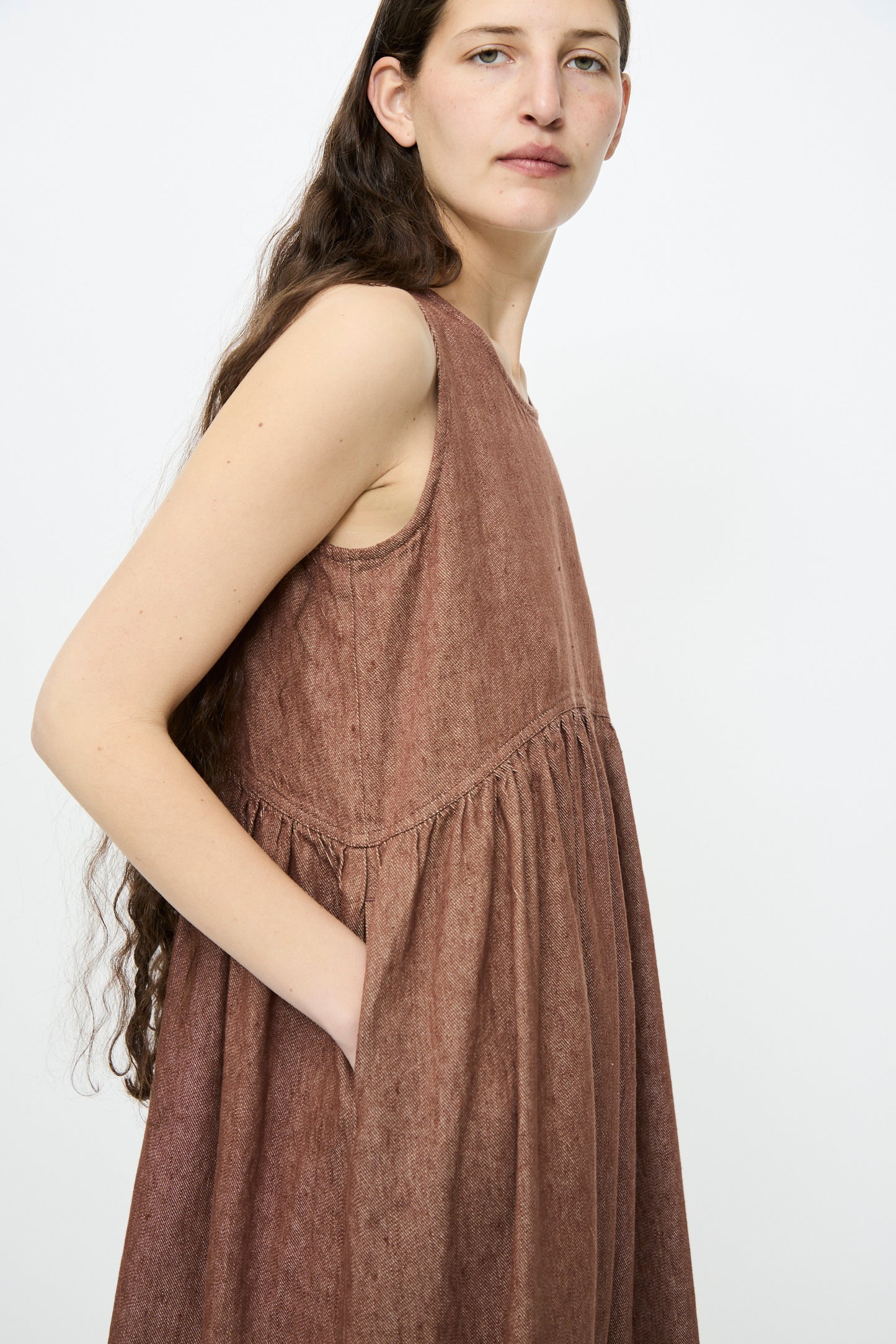 Against a white background, a person wears the Ichi Antiquités Linen Cotton Denim Dress in Brown. With long hair flowing and a hand in the pocket, looking forward.