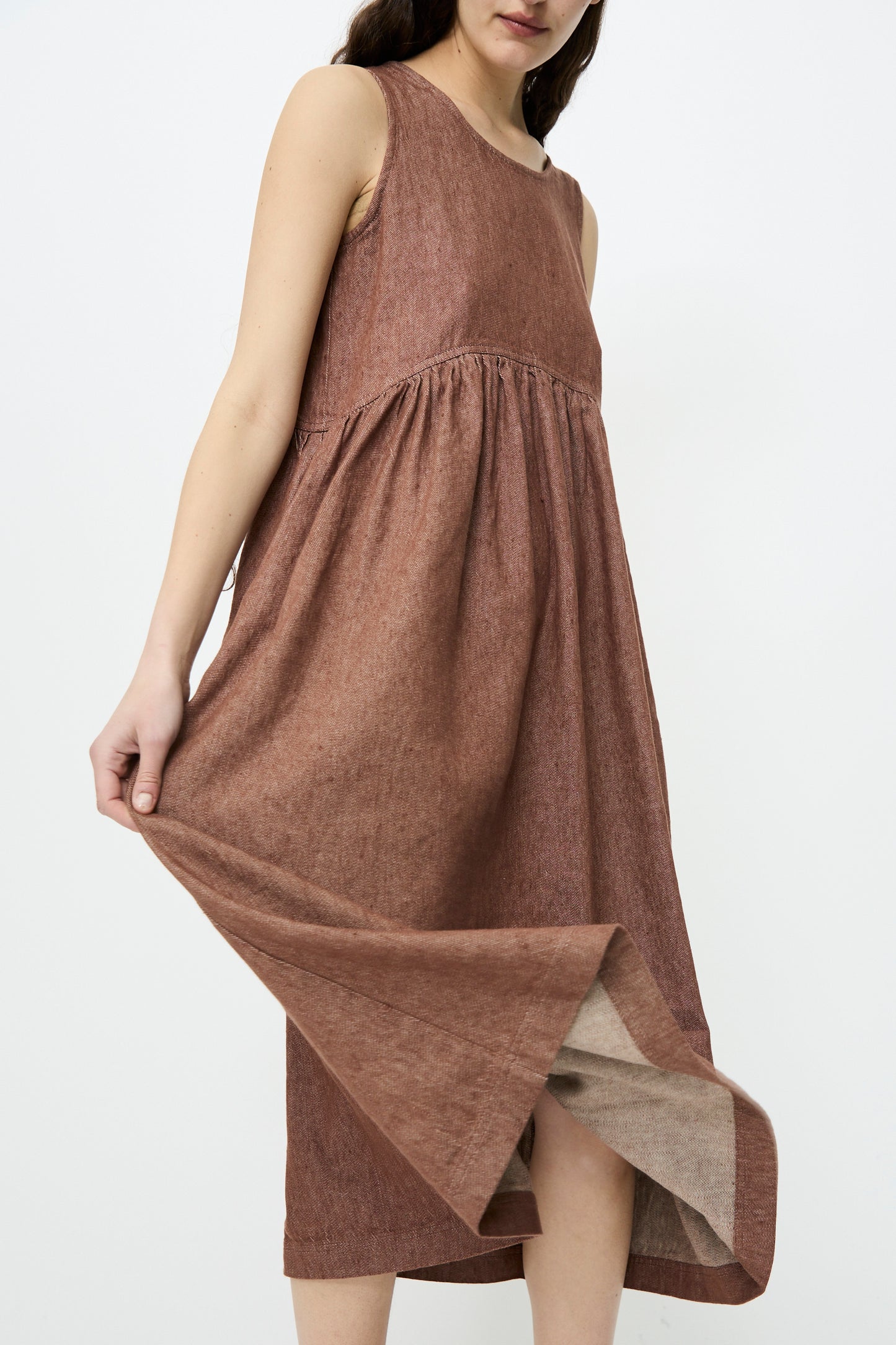 A person in the Ichi Antiquités Linen Cotton Denim Dress in brown, a sleeveless, ankle-length garment with a relaxed fit, delicately holding the skirt against a plain white backdrop.