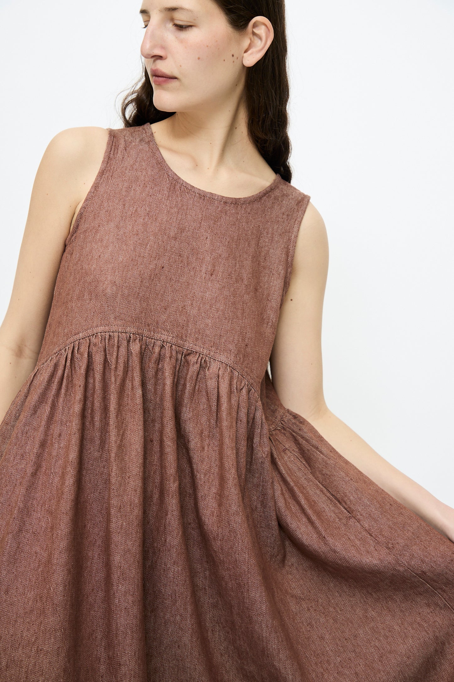 A person in the Ichi Antiquités Linen Cotton Denim Dress in brown, featuring a relaxed-fit and sleeveless design, gazes off to the side against a plain background.