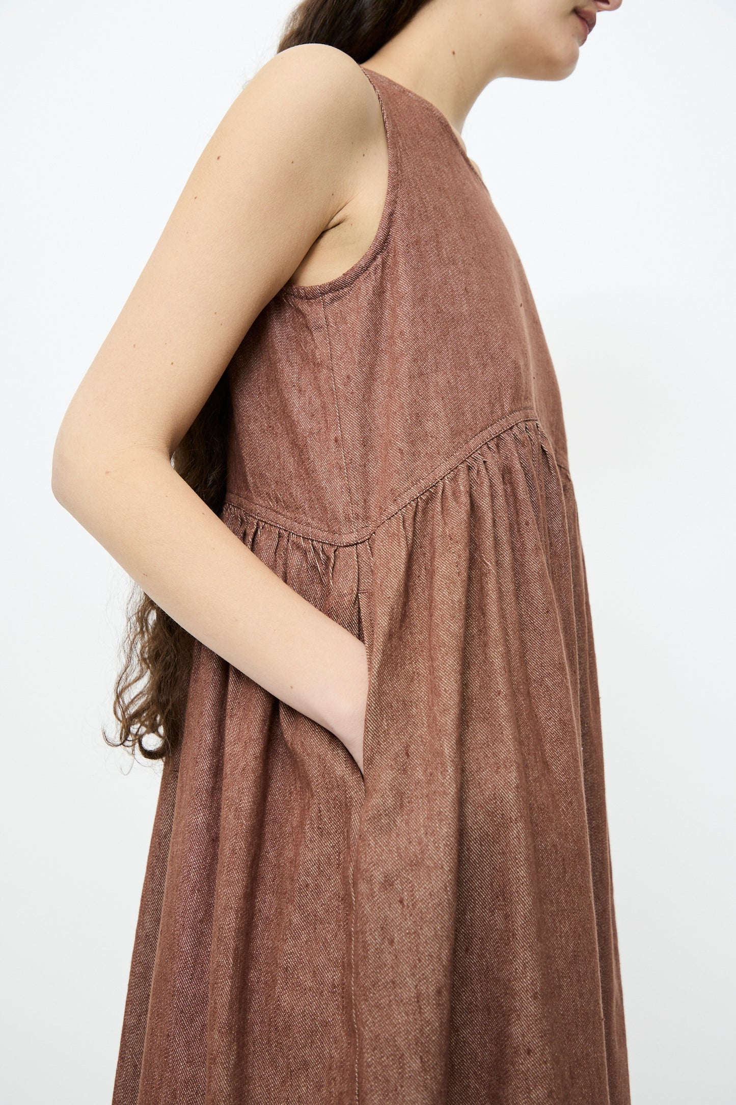 A person wearing the Linen Cotton Denim Dress from Ichi Antiquités, featuring a sleeveless design, relaxed fit, and brown color with one hand in a side pocket.