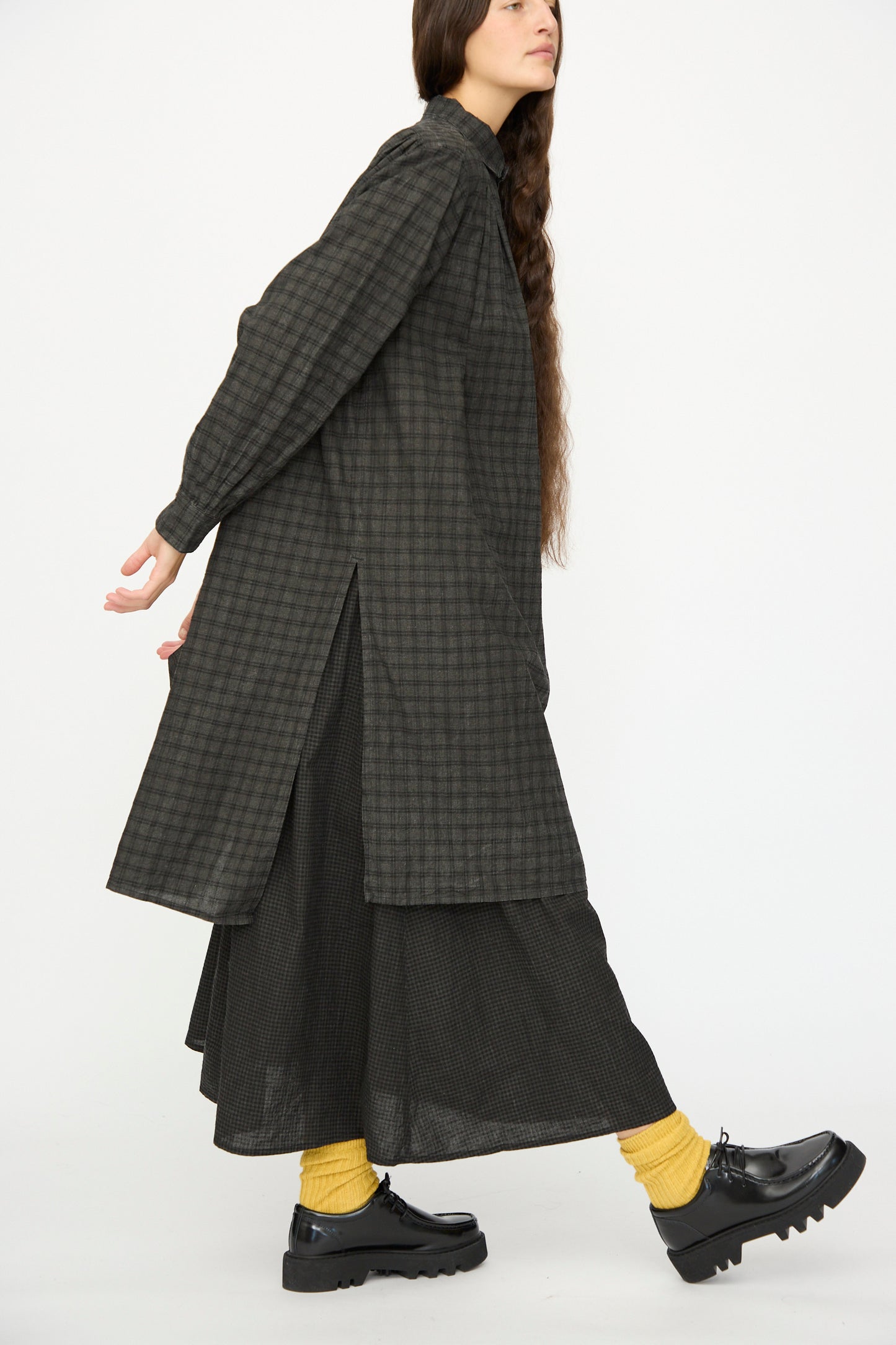 A person wearing the Ichi Antiquités Linen Cotton Gingham Sumi Shirt in Pin Check over a skirt, paired with yellow socks and black shoes, walks against a plain white background.
