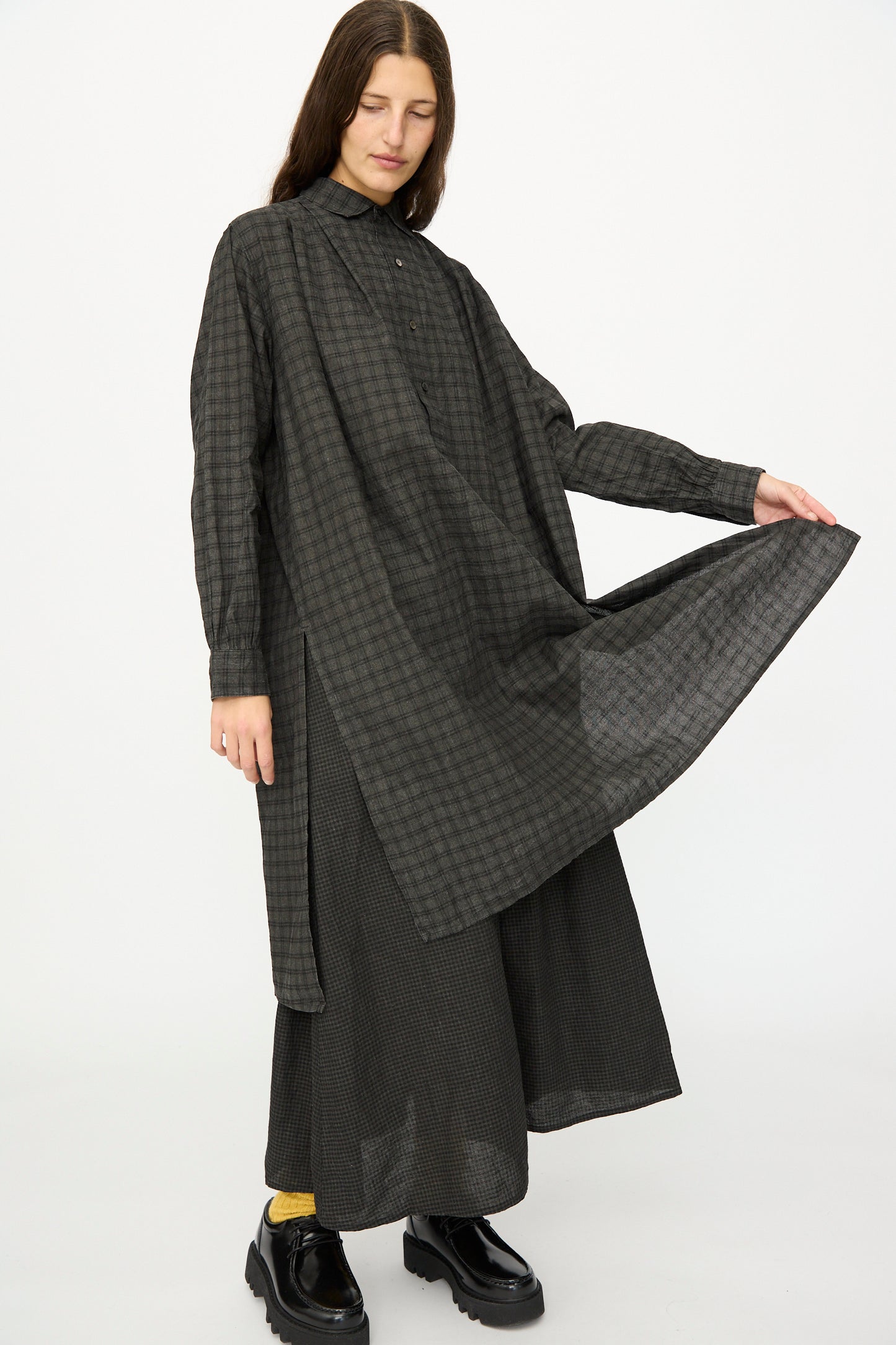 A person stands against a white background wearing the Ichi Antiquités Linen Cotton Gingham Sumi Shirt in Pin Check over wide pants and black shoes, holding out one side of the shirt.