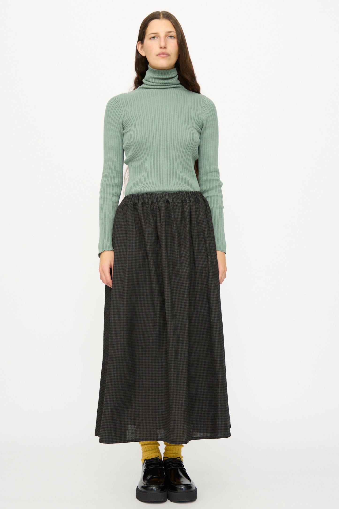 A woman stands facing forward against a white background, wearing a ribbed green turtleneck, yellow socks, black shoes, and the Ichi Antiquités Linen Cotton Gingham Sumi Skirt in Check.