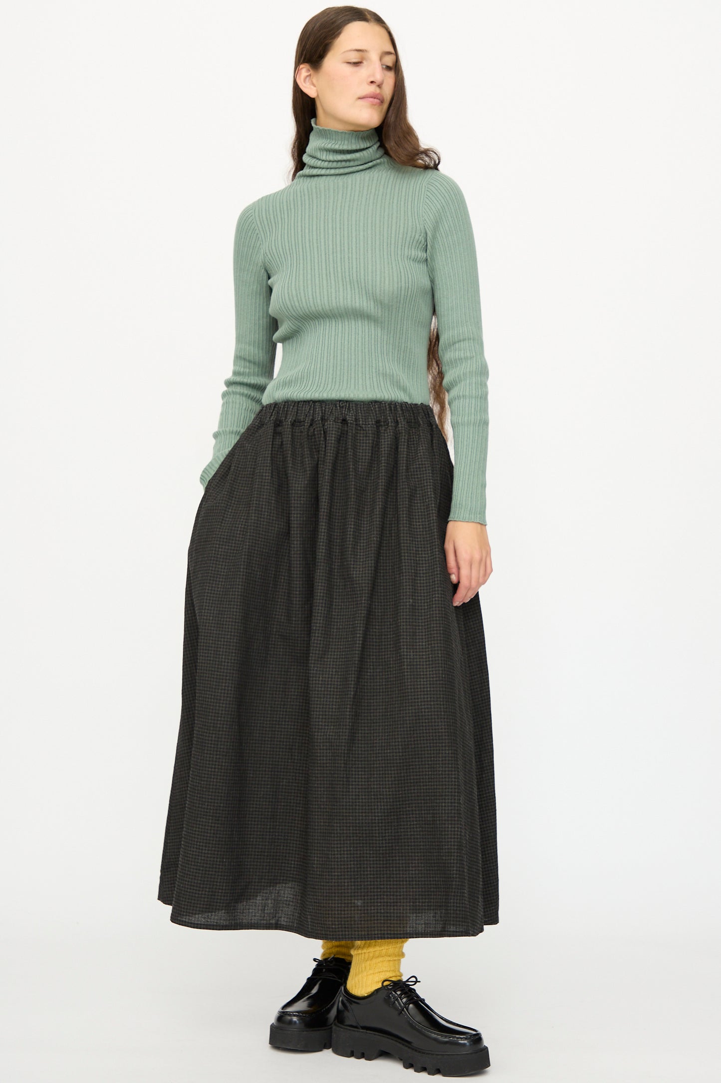 A woman stands against a plain white background wearing a green ribbed turtleneck, yellow socks, black shoes, and the Ichi Antiquités Linen Cotton Gingham Sumi Skirt in Check.