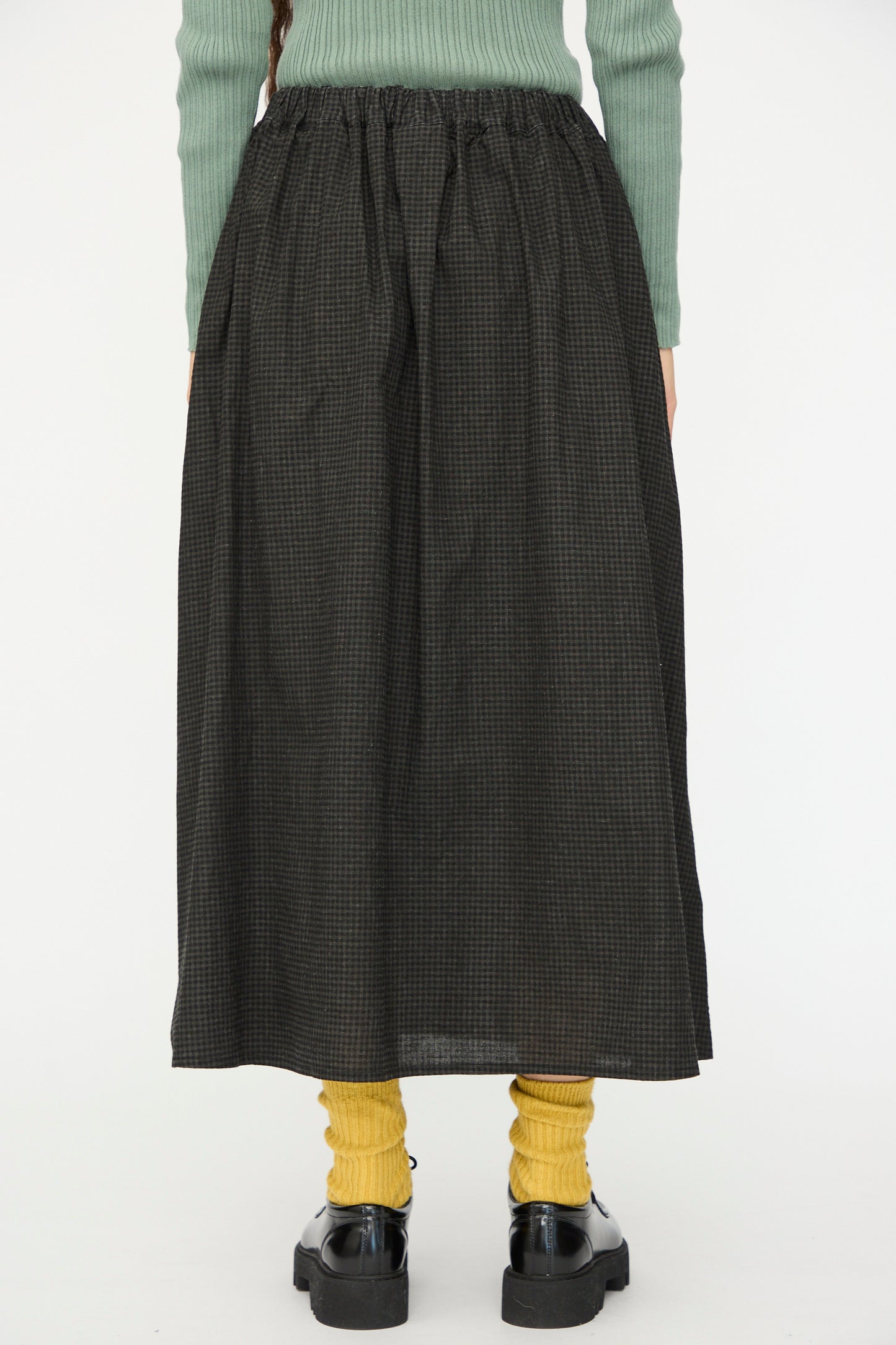 Someone stands against a plain white background wearing a green sweater, yellow socks, black shoes, and the Ichi Antiquités Linen Cotton Gingham Sumi Skirt in Check.