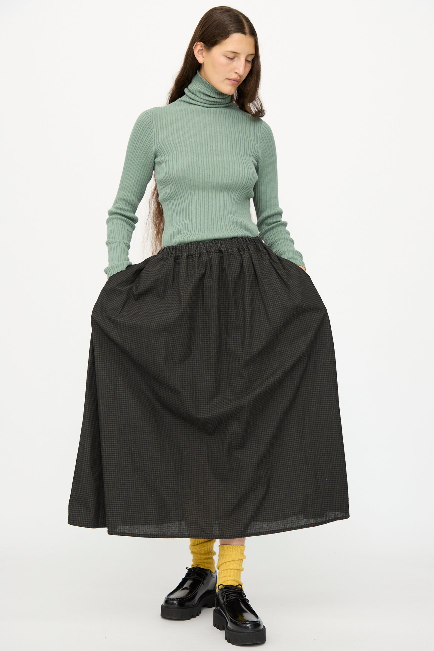 A woman wearing a light green turtleneck sweater, yellow socks, black shoes, and the Ichi Antiquités Linen Cotton Gingham Sumi Skirt in Check stands against a plain white background.