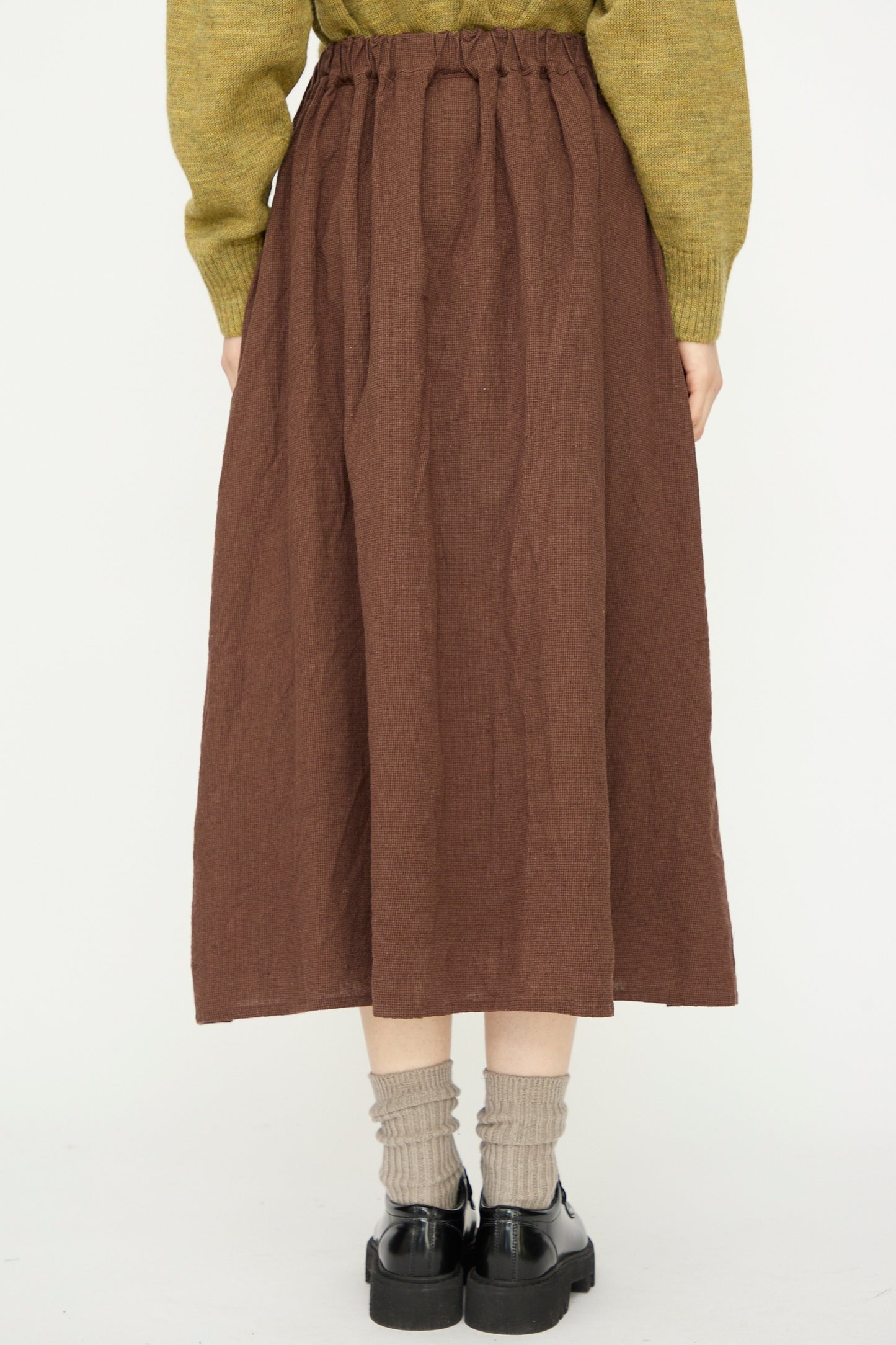 Person wearing the Ichi Antiquités Linen Cotton Houndstooth Skirt in Brown with a green sweater, beige socks, and black shoes, standing against a plain white background.