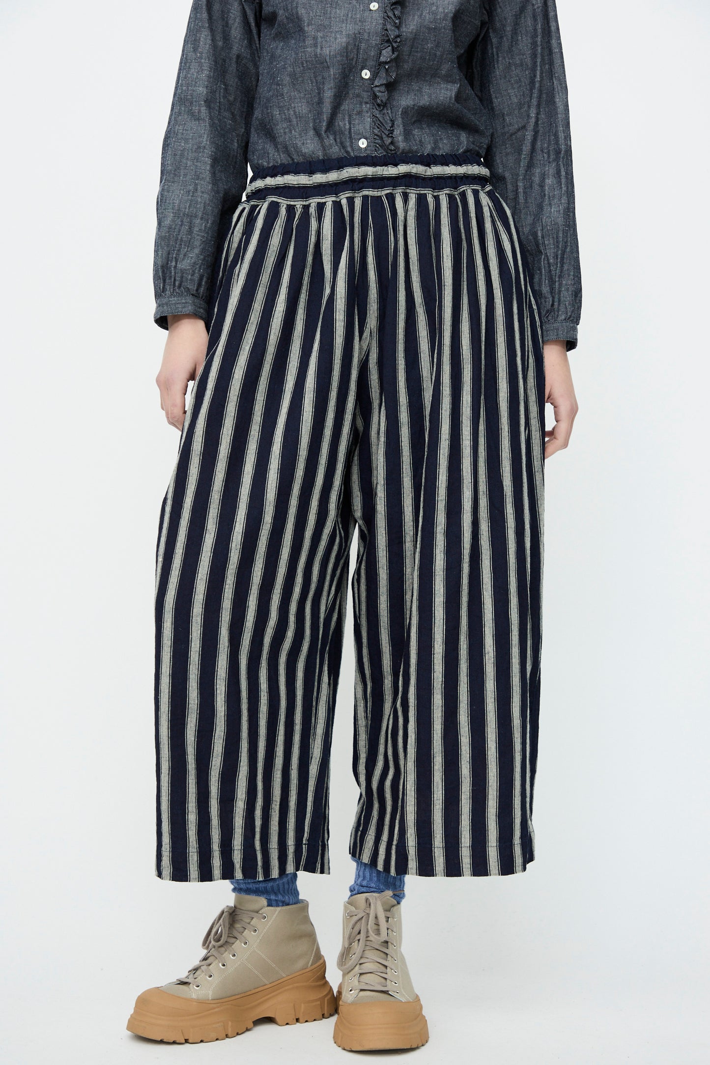 A person wearing a dark button-up shirt, Ichi Antiquités' Linen Cotton Stripe Pants in Indigo and Natural with an elastic waist, and beige high-top sneakers stands against a plain background.