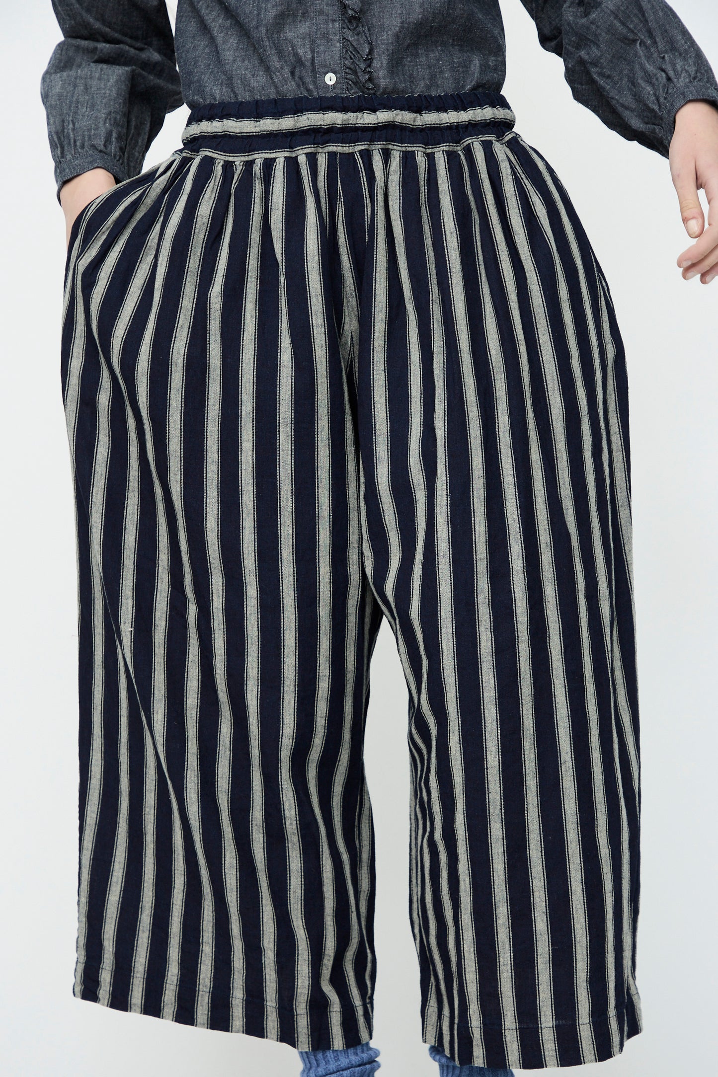 A person wears Linen Cotton Stripe Pants by Ichi Antiquités, featuring vertical indigo and natural stripes and an elasticated waist. Their hands rest casually in the pockets of these stylish wide-legged trousers.