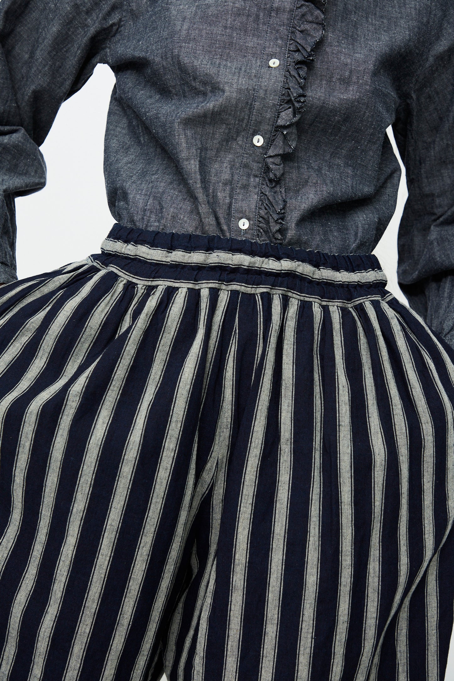 A person is wearing a dark button-up shirt with ruffled details and Ichi Antiquités Linen Cotton Stripe Pants in indigo and natural, which are high-waisted and cropped with a wide-leg design.