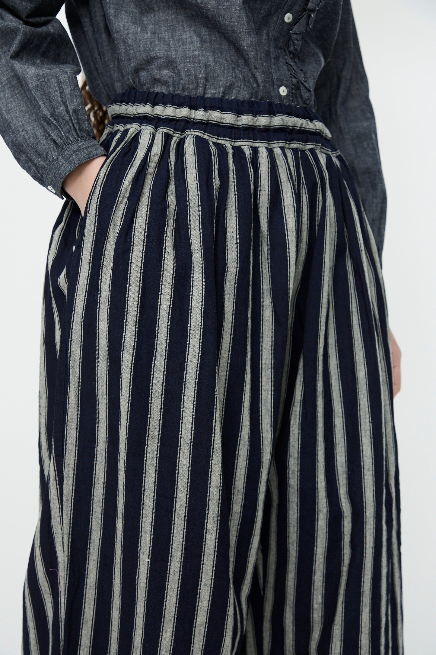 Person wearing a gray button-up shirt tucked into Linen Cotton Stripe Pants in Indigo and Natural by Ichi Antiquités, featuring high-waisted, navy and white vertical stripes with an elasticated waist.