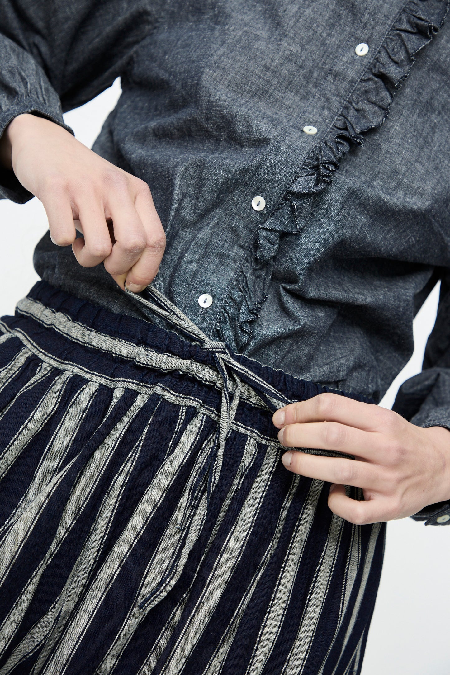 Wearing a ruffled button-up shirt, an individual adjusts the drawstring on Ichi Antiquités Linen Cotton Stripe Pants in Indigo and Natural with an elasticated waist.