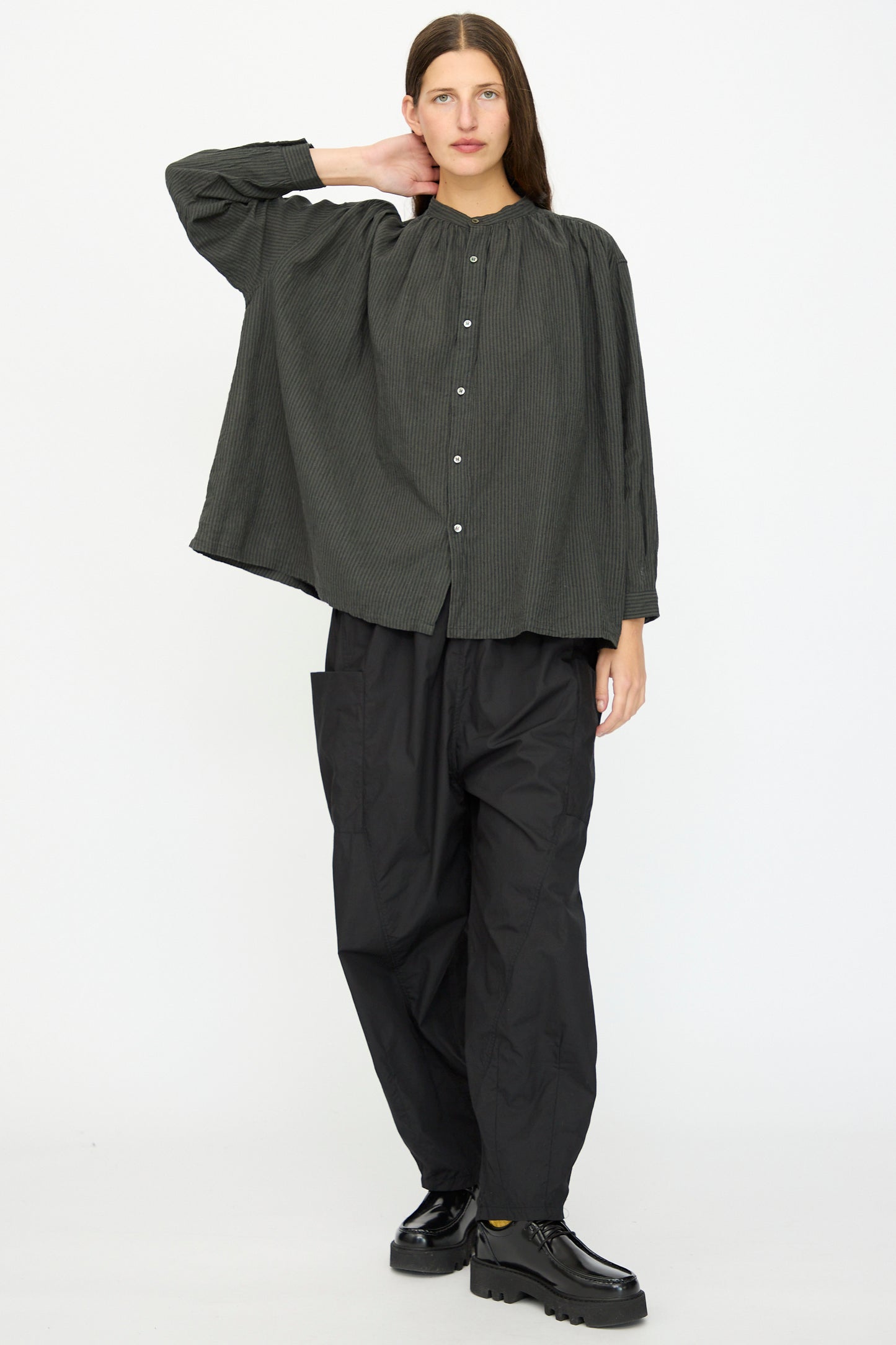A person stands against a plain background wearing the Ichi Antiquités Linen and Cotton Stripe Shirt in Charcoal, paired with baggy black pants and black shoes, one hand resting on the back of their neck.