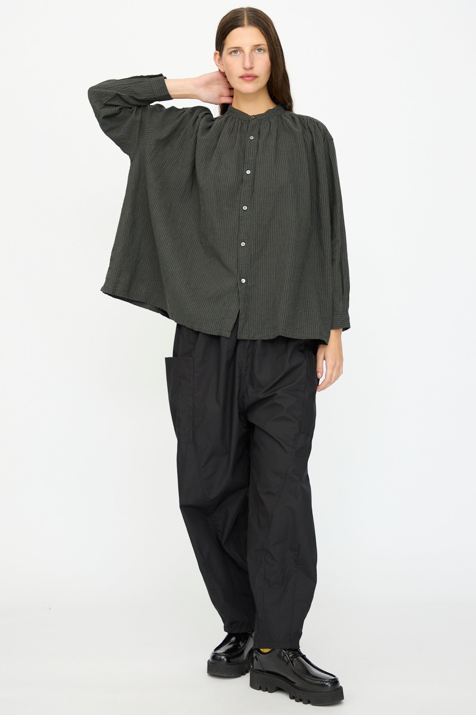 A person stands against a plain background wearing the Ichi Antiquités Linen and Cotton Stripe Shirt in Charcoal, paired with baggy black pants and black shoes, one hand resting on the back of their neck.