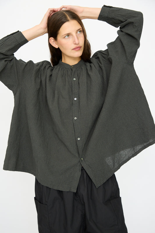 A person wears the Ichi Antiquités Linen and Cotton Stripe Shirt in Charcoal, styled oversized with black pants, standing against a plain background with hands resting on their head.