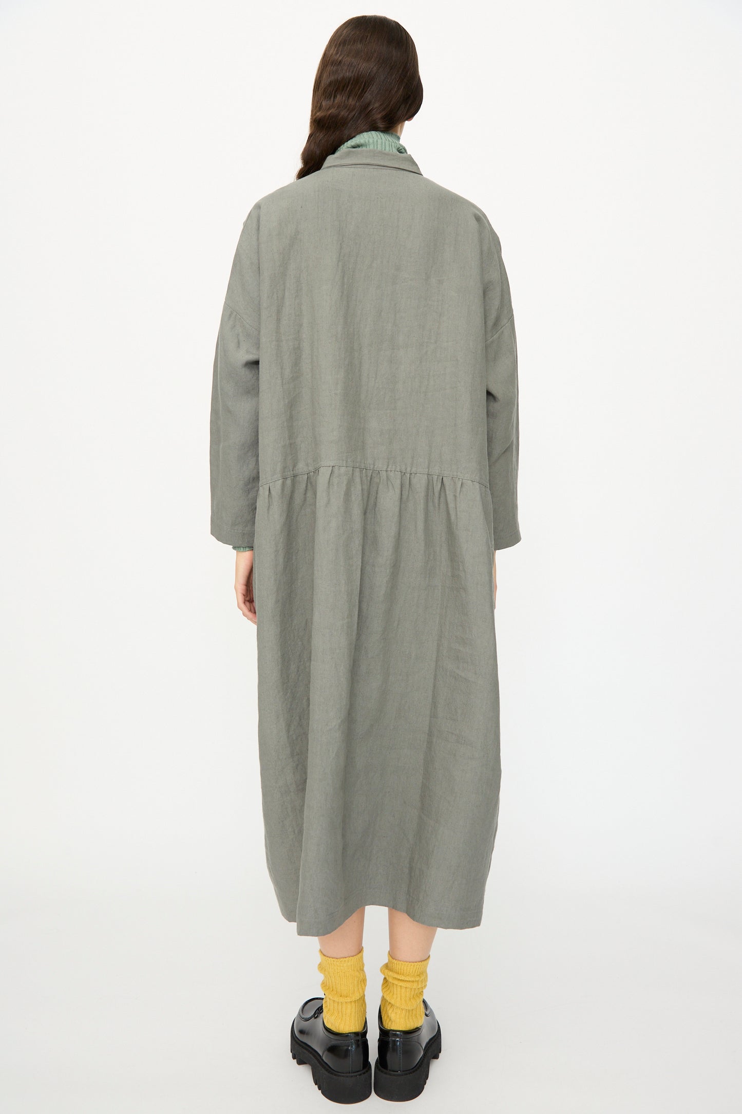 A person stands facing away, wearing the Ichi Antiquités Linen Dress in Khaki with yellow socks and black platform shoes against a plain white background. This elegant long sleeve, gathered waist dress is made in Japan.