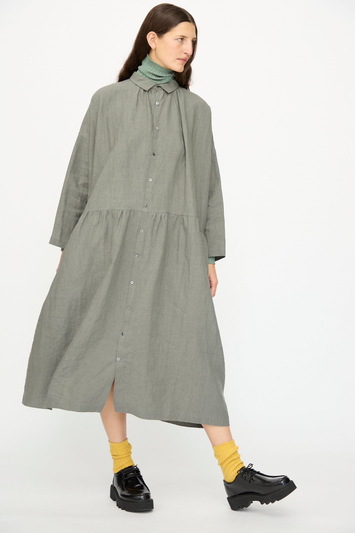 A woman stands against a plain white background wearing the Ichi Antiquités Linen Dress in Khaki, styled over a green turtleneck, with yellow socks and black platform shoes.