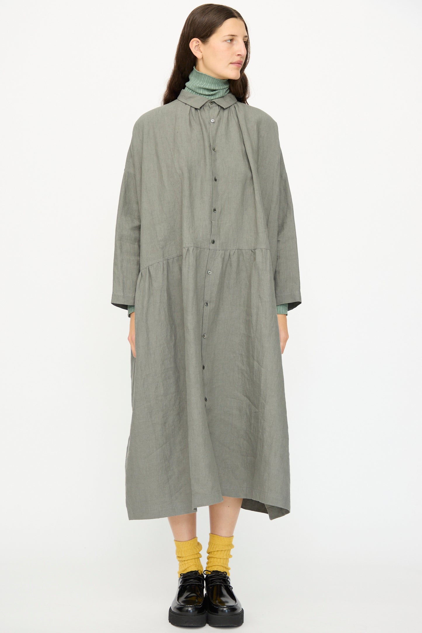 A woman wears the Ichi Antiquités Linen Dress in Khaki over a green turtleneck, paired with yellow socks and black shoes, standing against a plain white background.