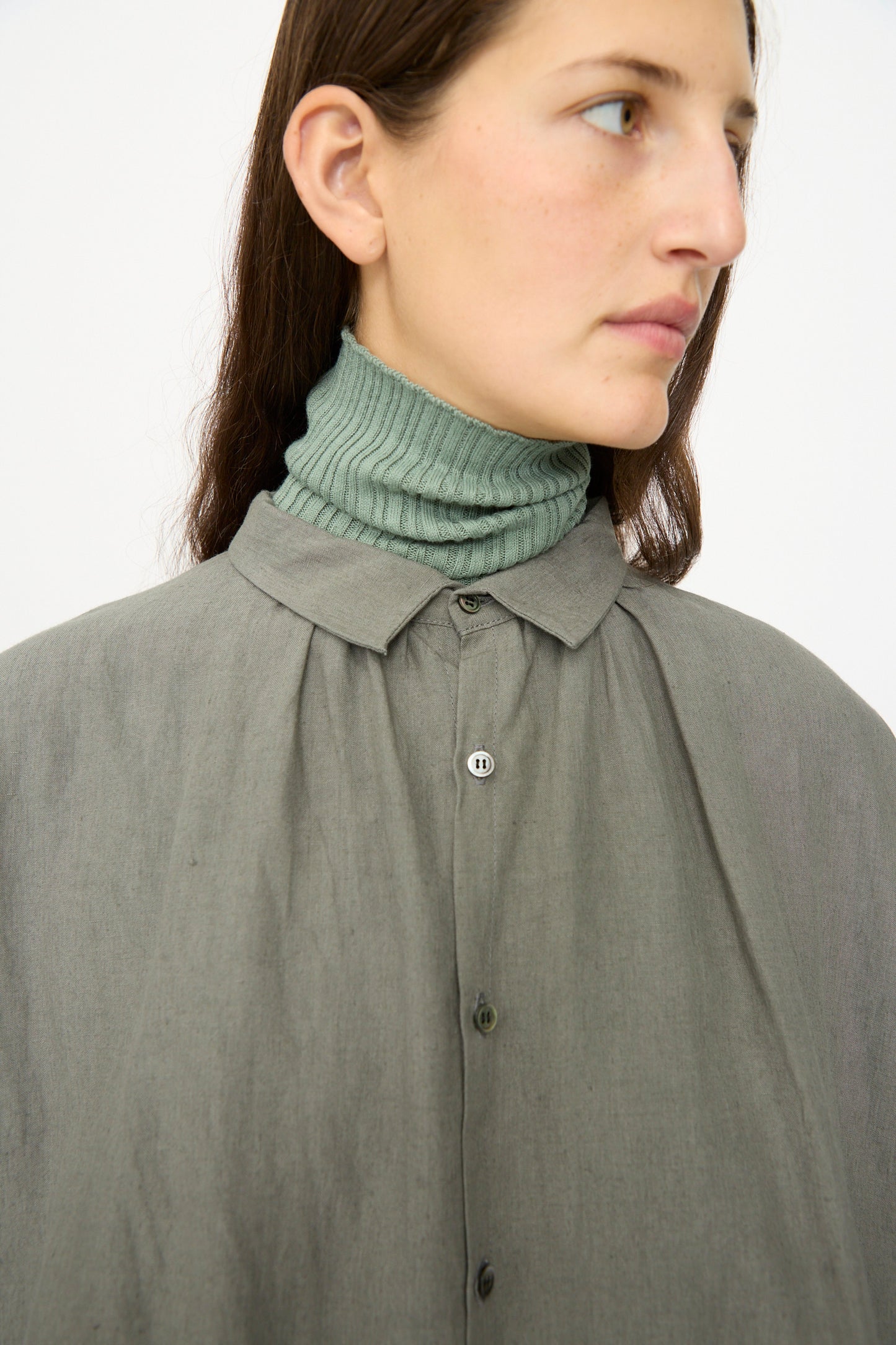 A person wears the Ichi Antiquités Linen Dress in Khaki, styled over a ribbed green turtleneck, gazing to the side against a plain background.
