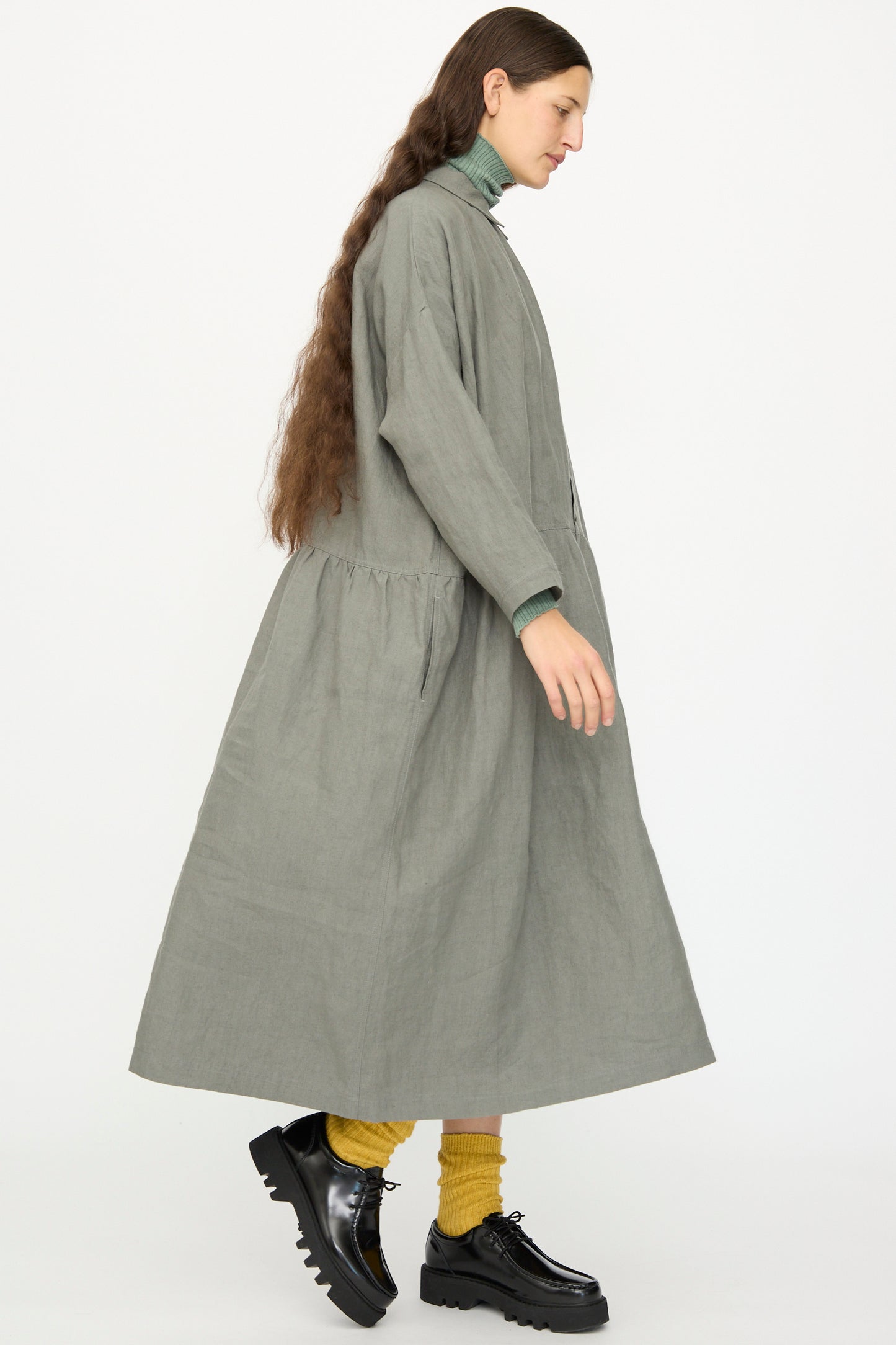 A person with long brown hair wears the Ichi Antiquités Linen Dress in Khaki, paired with a green turtleneck, yellow socks, and black platform shoes, walking against a plain white background.