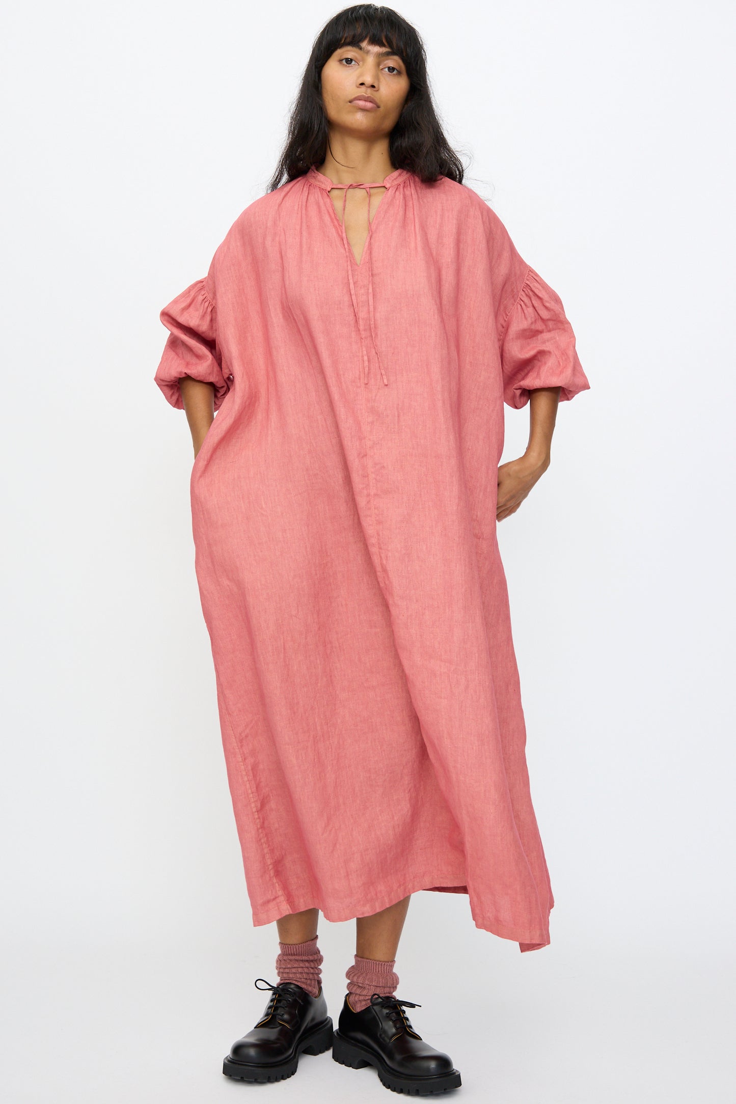 A woman stands against a plain background wearing the Ichi Antiquités Linen Dress in Pink, a loose-fitting, hand-dyed linen maxi dress with gathered sleeves, paired with black shoes and pink socks.