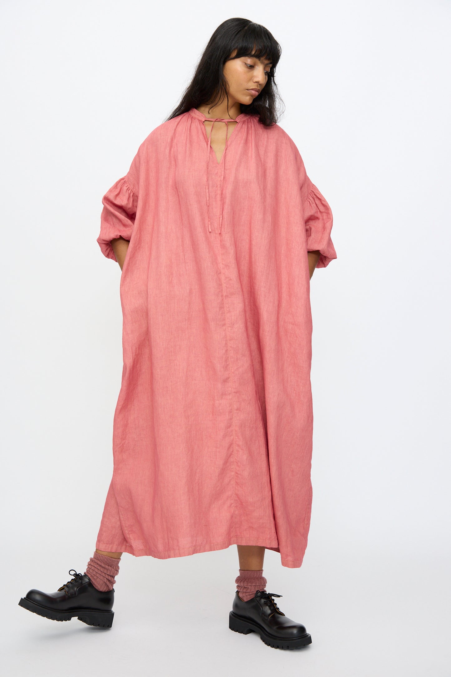 A woman stands against a white background wearing the Ichi Antiquités Linen Dress in Pink—an oversized, hand-dyed maxi with puffed sleeves—paired with black shoes and matching pink socks.