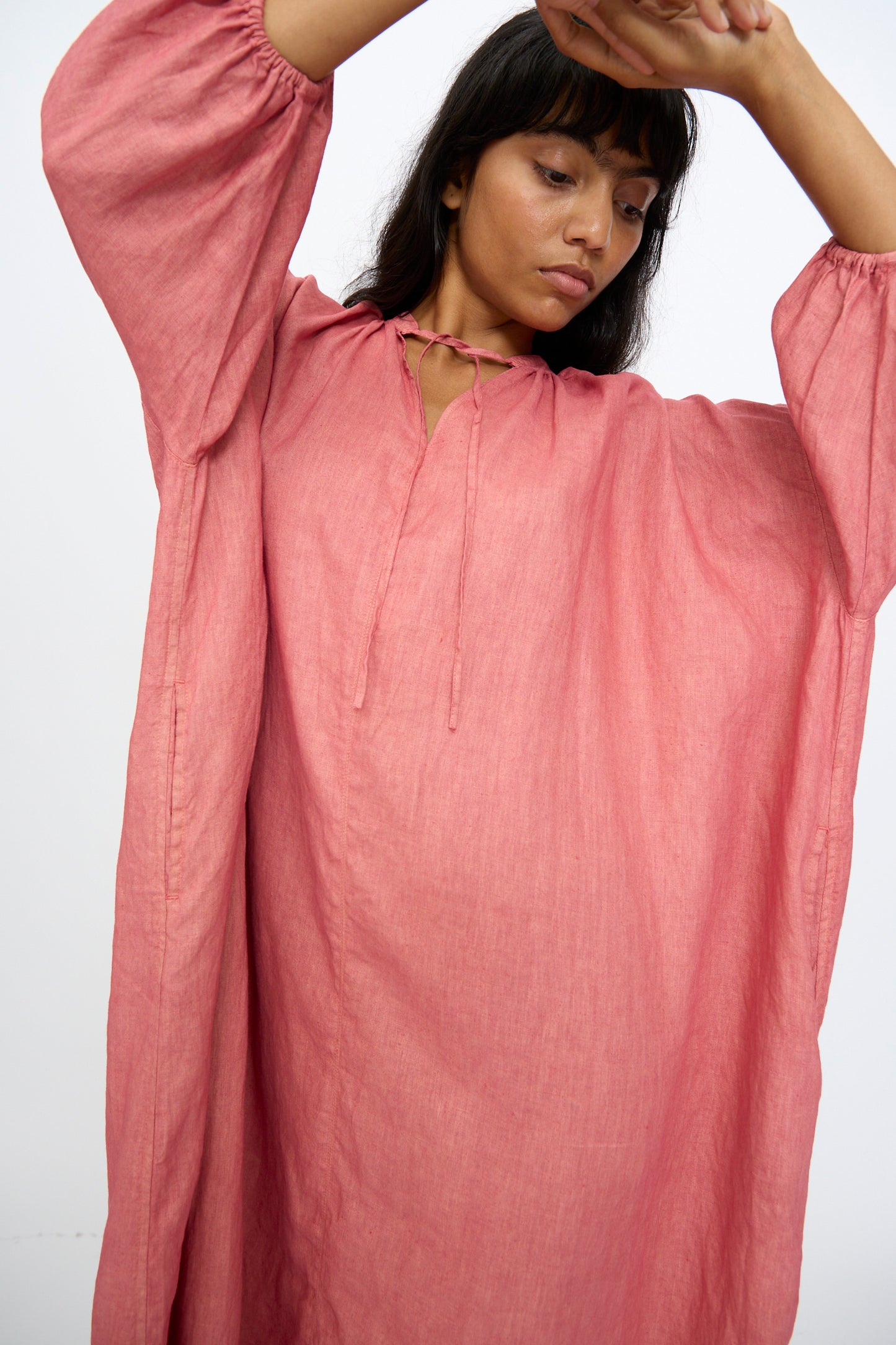 A person models the Ichi Antiquités Linen Dress in Pink, a loose-fitting, long-sleeved maxi dress, standing with arms raised against a plain light background.