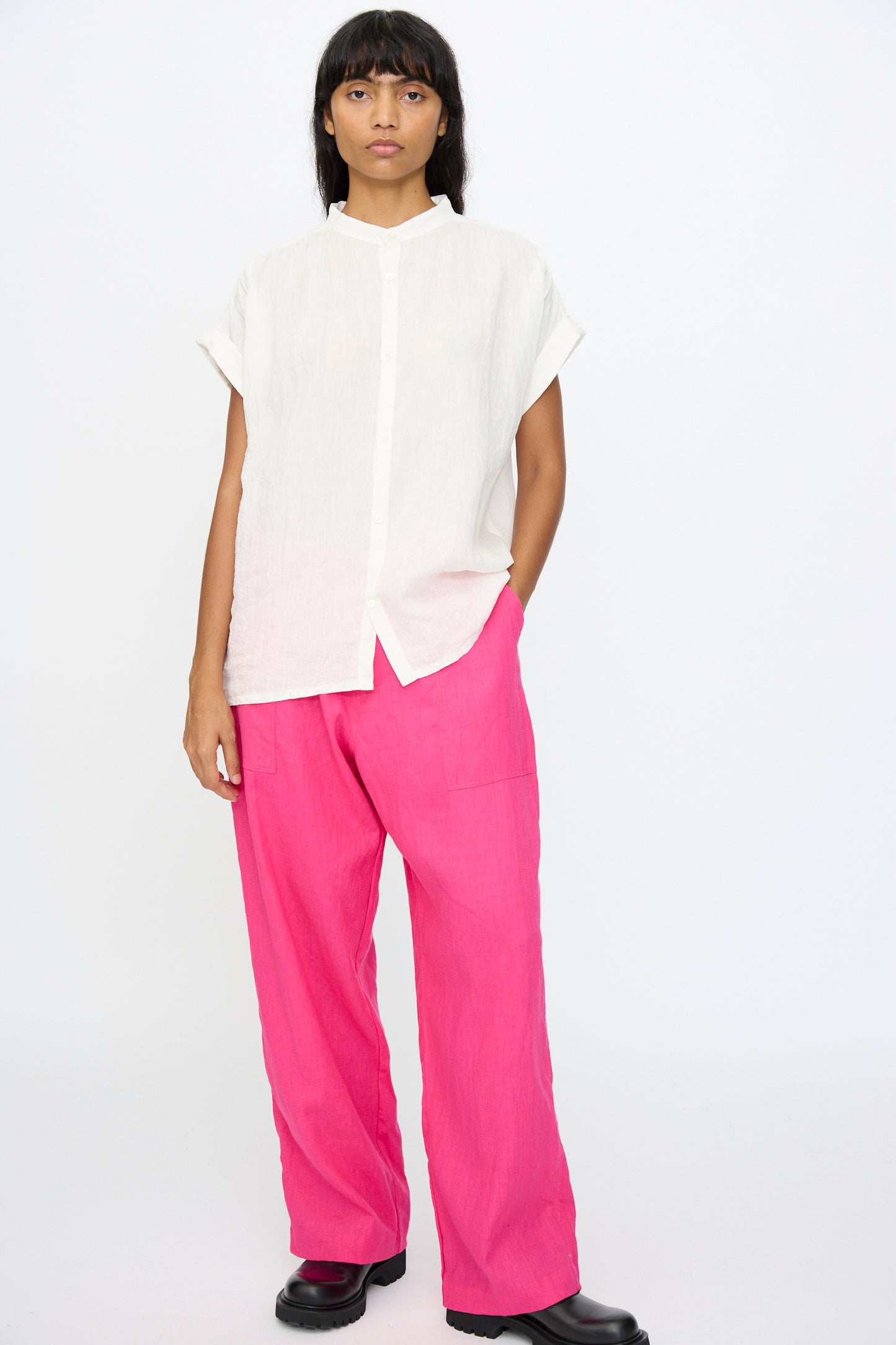 A person stands against a plain white background, wearing the Ichi Antiquités Linen Dungaree Blouse in White, bright pink wide-leg pants, and black shoes.