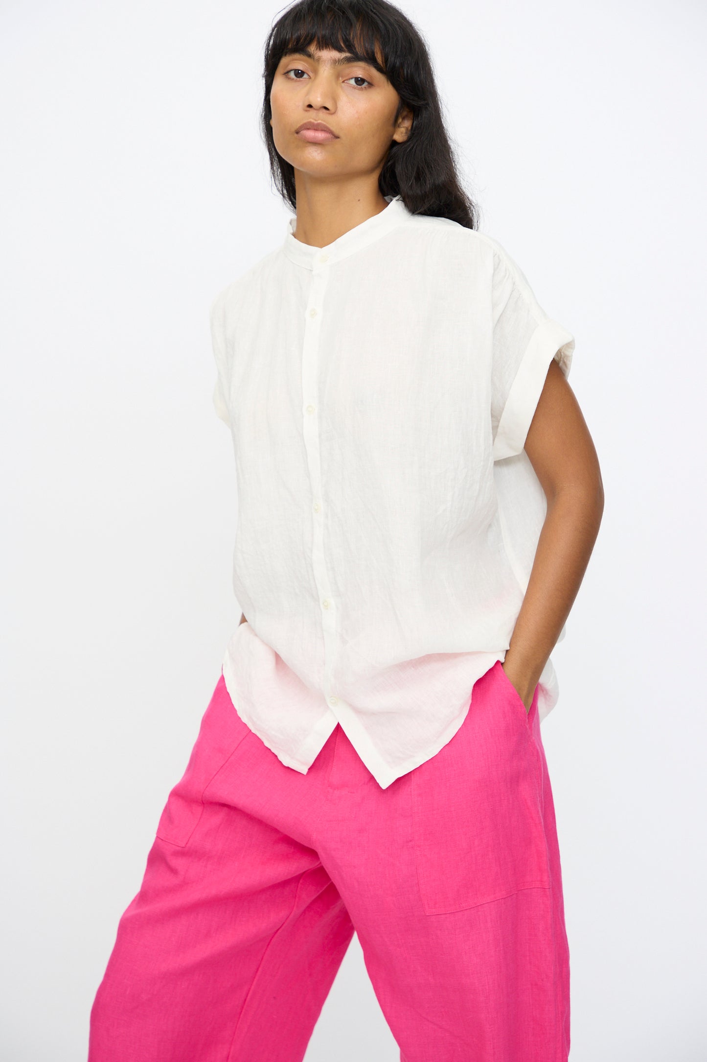 A person stands against a plain background wearing the Ichi Antiquités Linen Dungaree Blouse in White with loose-fitting bright pink pants, one hand in their pocket.