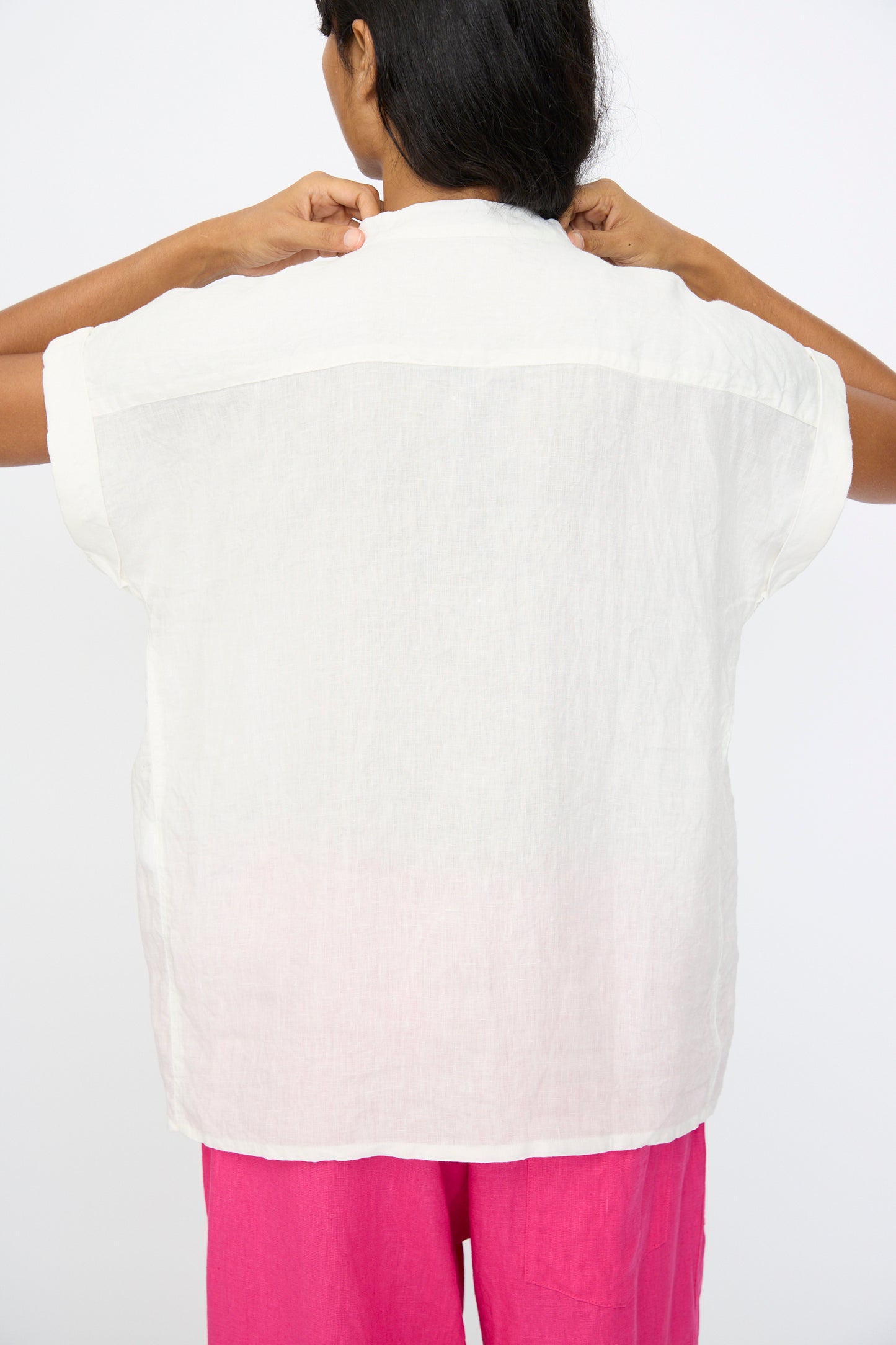A person faces away, adjusting the collar of the Ichi Antiquités Linen Dungaree Blouse in White with short sleeves, paired with bright pink pants against a plain background.