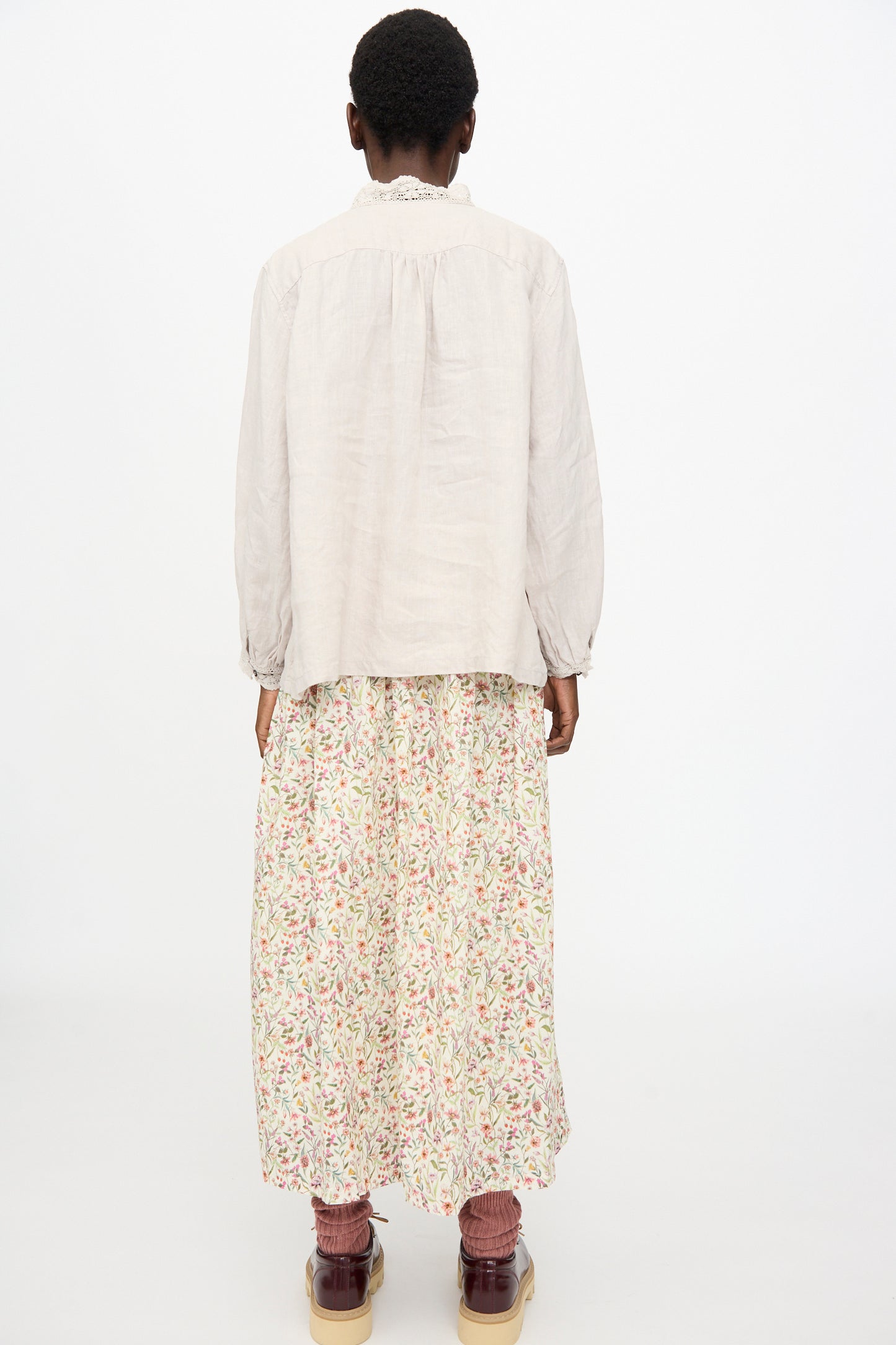 A person stands with their back to the camera, wearing the Ichi Antiquités Linen Shirt in Greige, an oversized fit style, paired with a floral ankle-length skirt, maroon socks, and brown shoes against a plain white background.