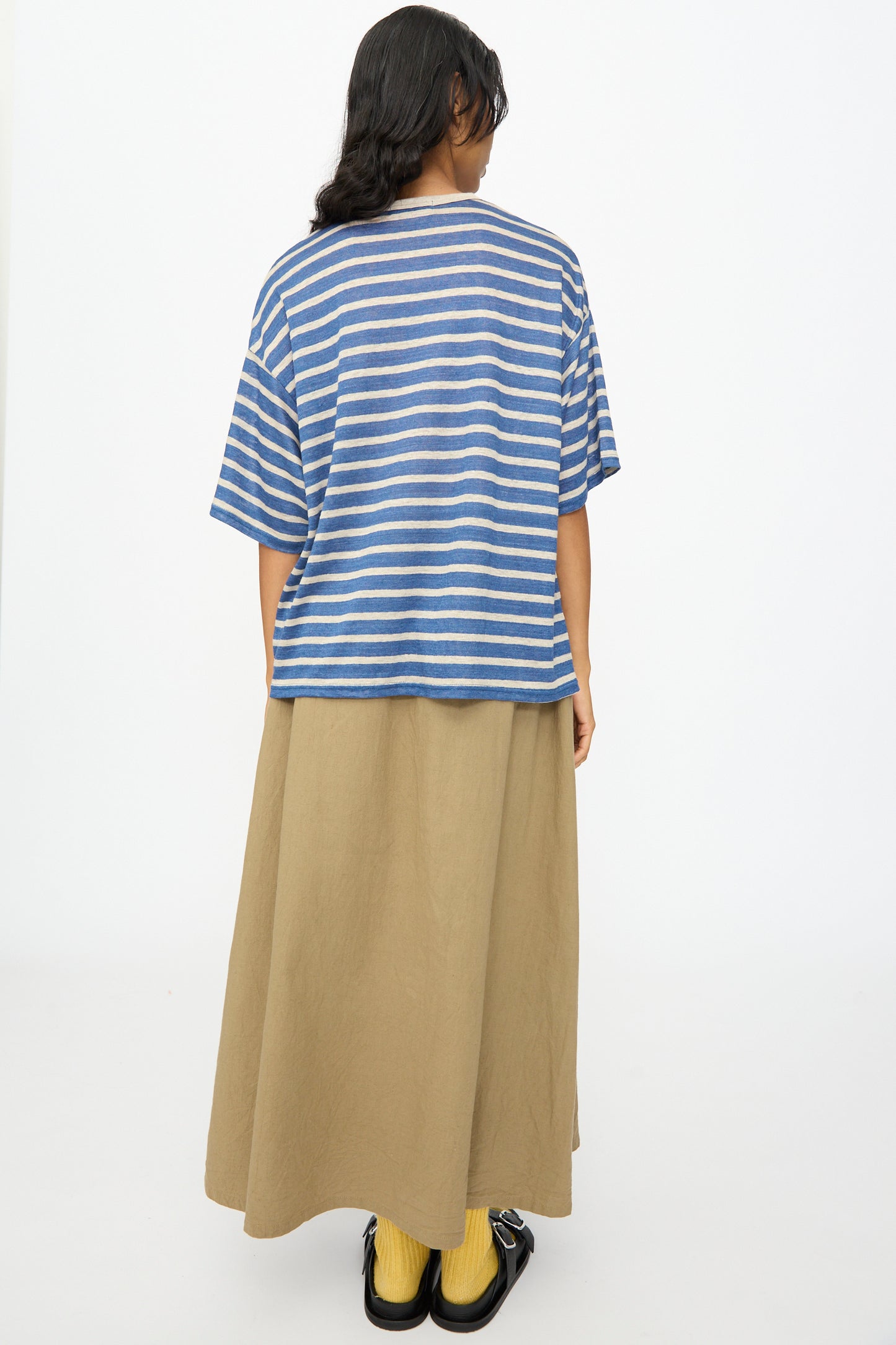 A person stands facing away against a white background, wearing the Ichi Antiquités Linen Stripe Pullover in Blue and Natural, paired with a long tan skirt, yellow socks, and black shoes.