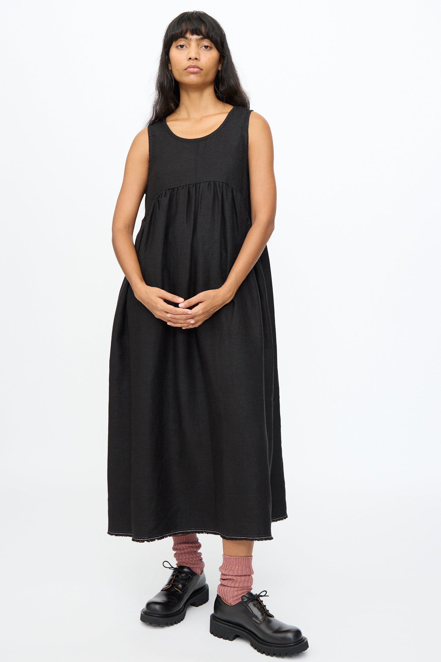 A person stands against a plain background wearing the Ichi Antiquités Twill Weave Linen Dress in Black, paired with pink socks and black lace-up shoes.