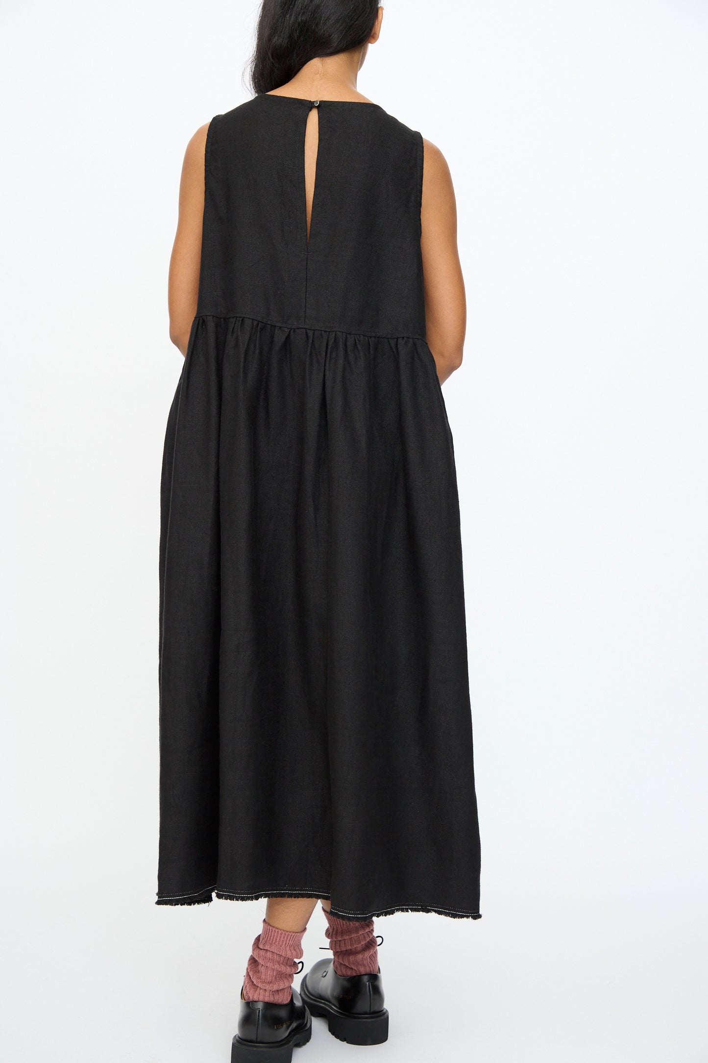 A person stands facing away, wearing the Ichi Antiquités Twill Weave Linen Dress in Black featuring a sleeveless design and back keyhole opening, paired with black shoes and pink socks.