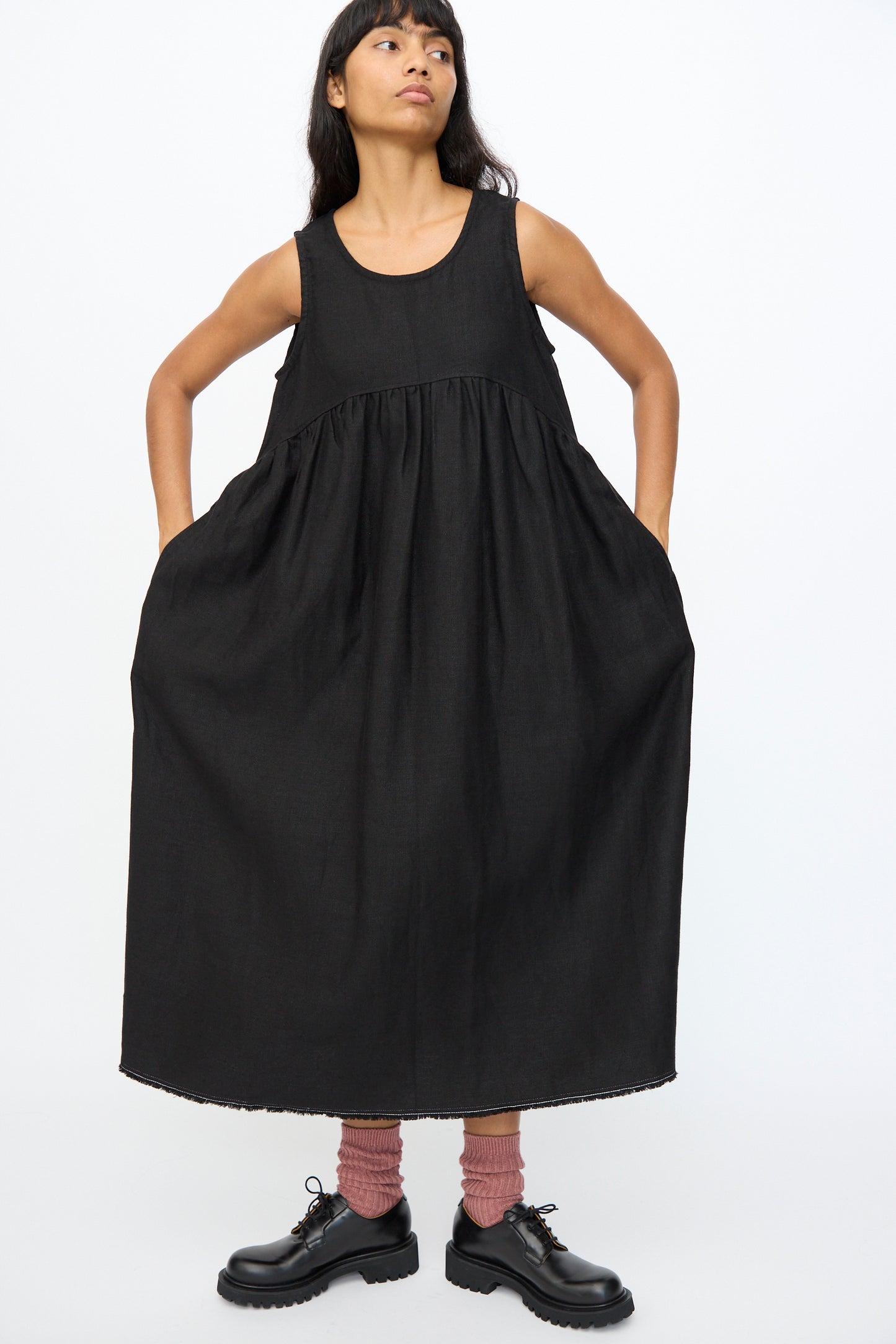 A person stands against a white background wearing the Ichi Antiquités Twill Weave Linen Dress in Black, paired with pink socks and black shoes, hands tucked into the dress's side pockets.
