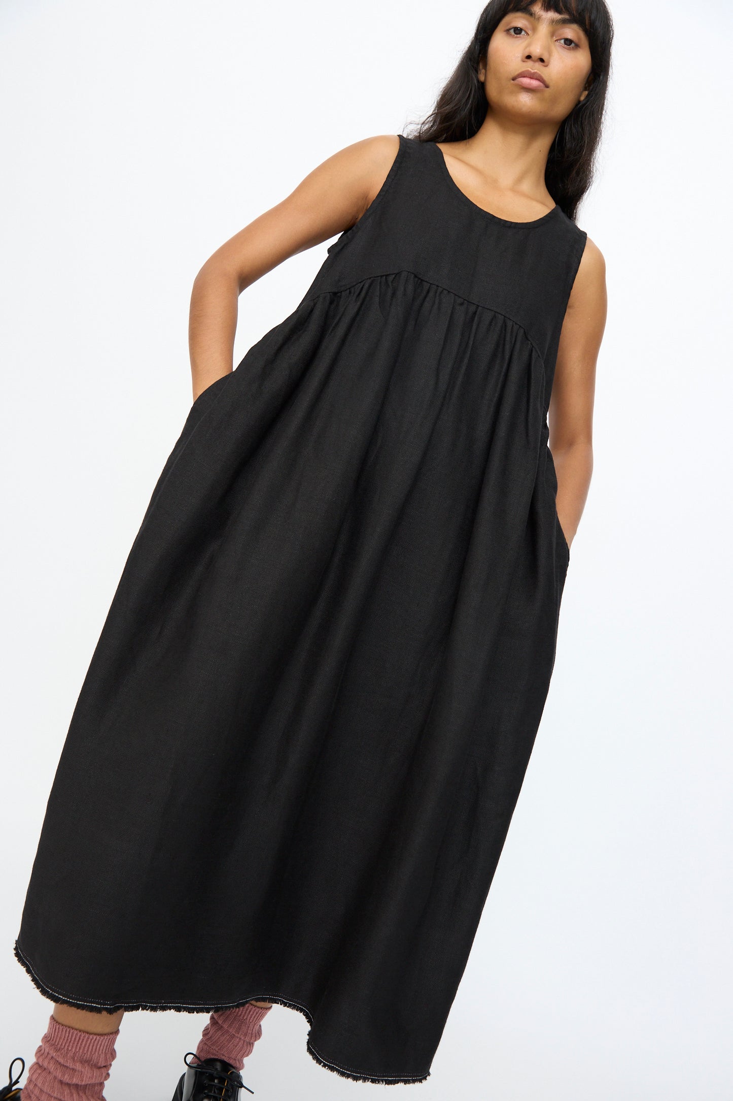A woman wearing the Ichi Antiquités Twill Weave Linen Dress in Black, paired with pink socks and black shoes, stands against a plain white background.