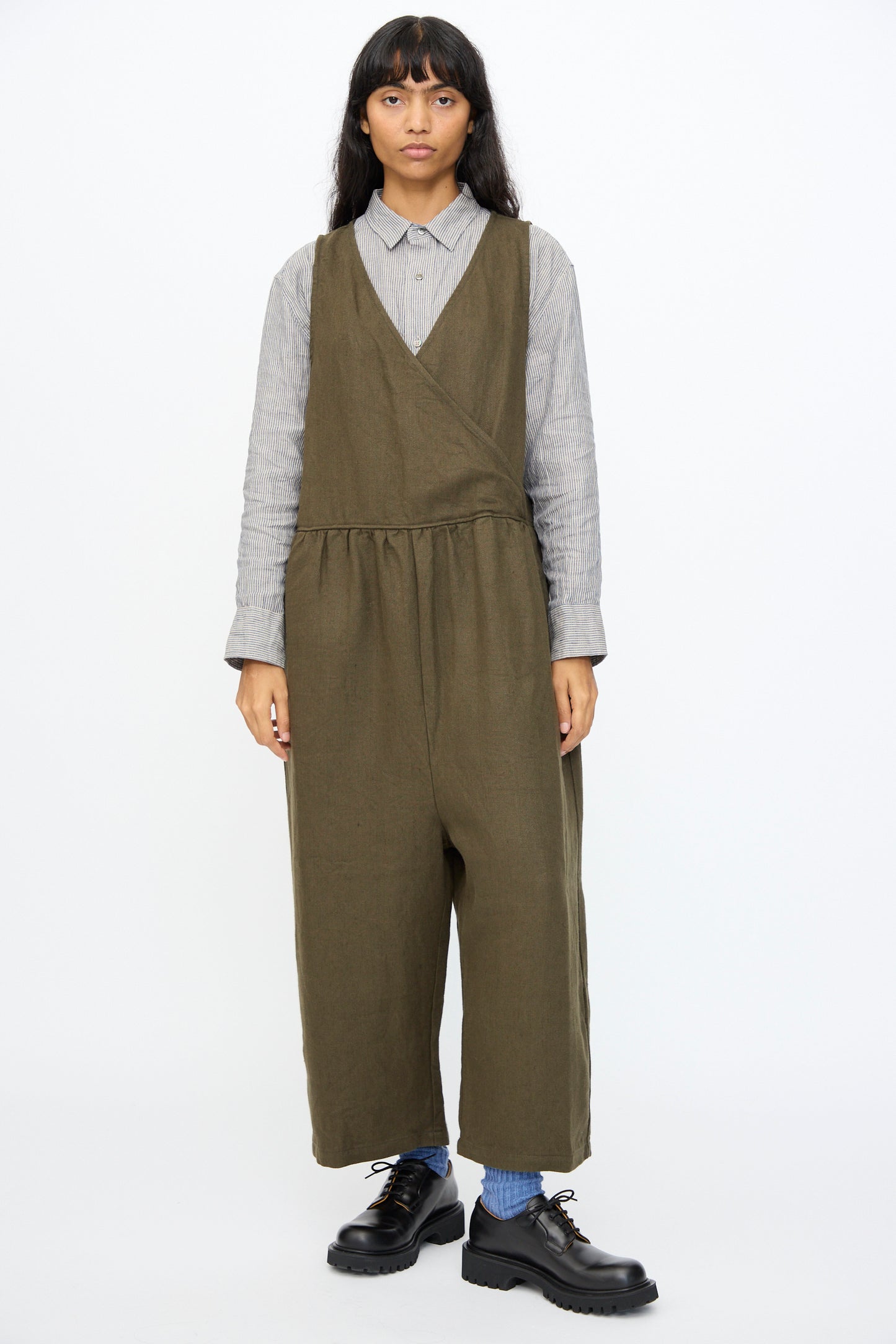 Person wearing the Ichi Antiquités Twill Weave Linen Jumpsuit in Khaki over a gray collared shirt, paired with blue socks and black shoes, standing against a plain background.
