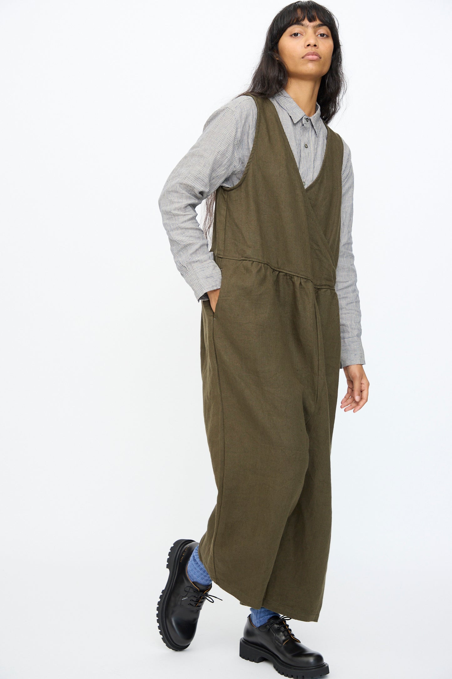 A person wears the Ichi Antiquités Twill Weave Linen Jumpsuit in Khaki over a gray button-up shirt, blue socks, and black shoes, standing against a plain white background.