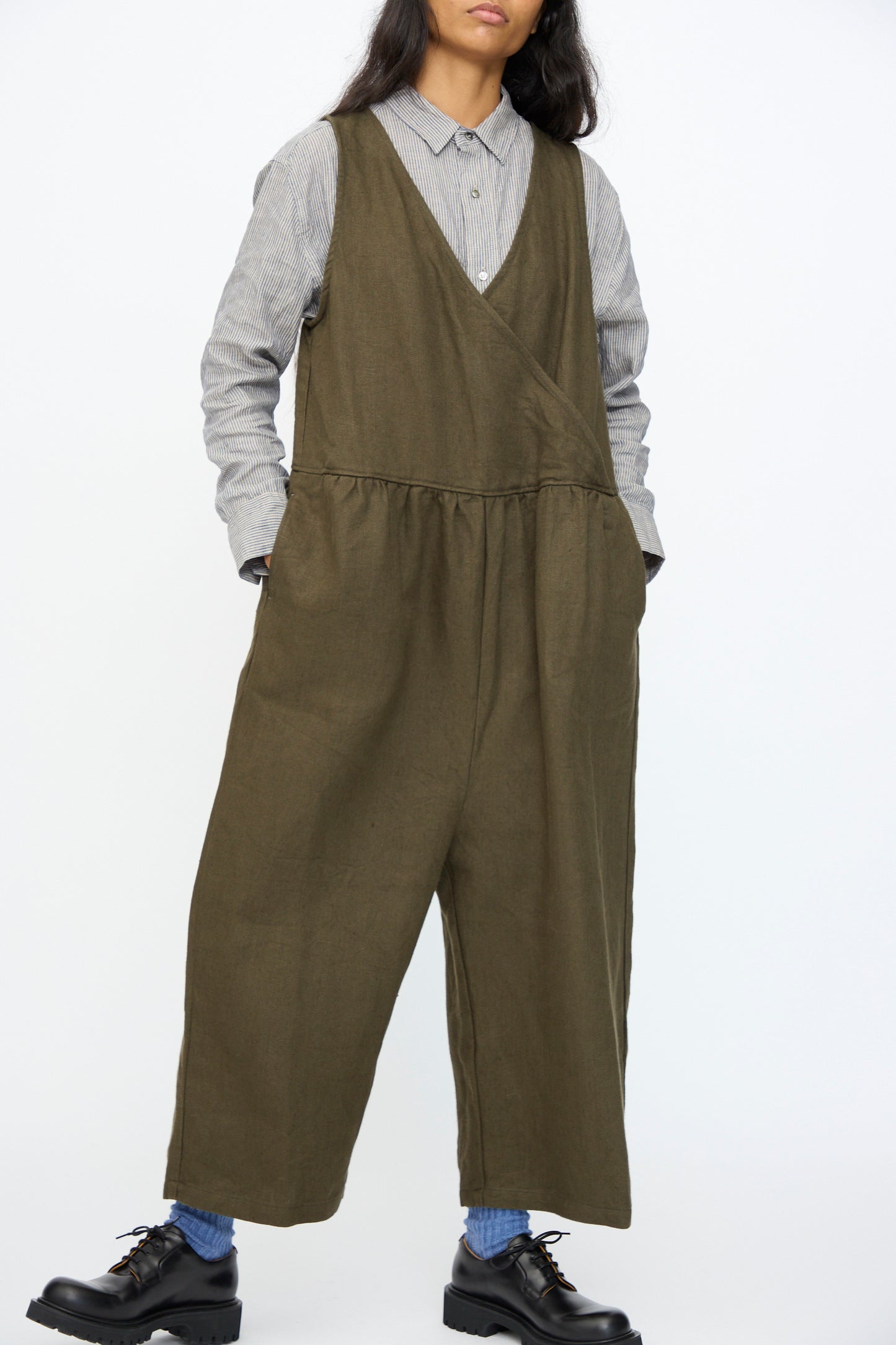 A person wears the Ichi Antiquités Twill Weave Linen Jumpsuit in Khaki over a gray button-up shirt, paired with black shoes and blue socks, standing against a plain white background.