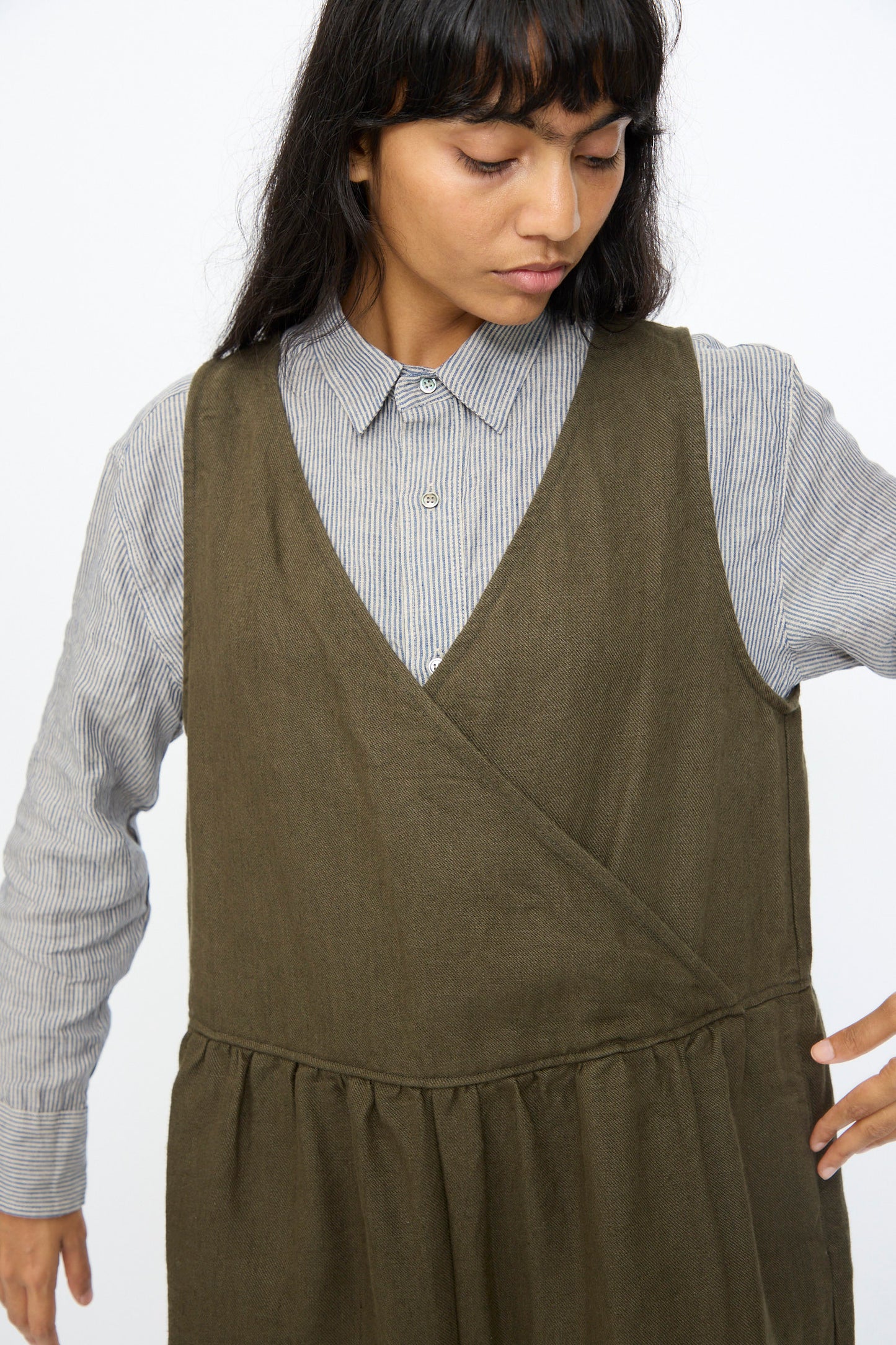 A person wears the Ichi Antiquités Twill Weave Linen Jumpsuit in Khaki over a striped button-up shirt, looking down with one arm slightly raised.
