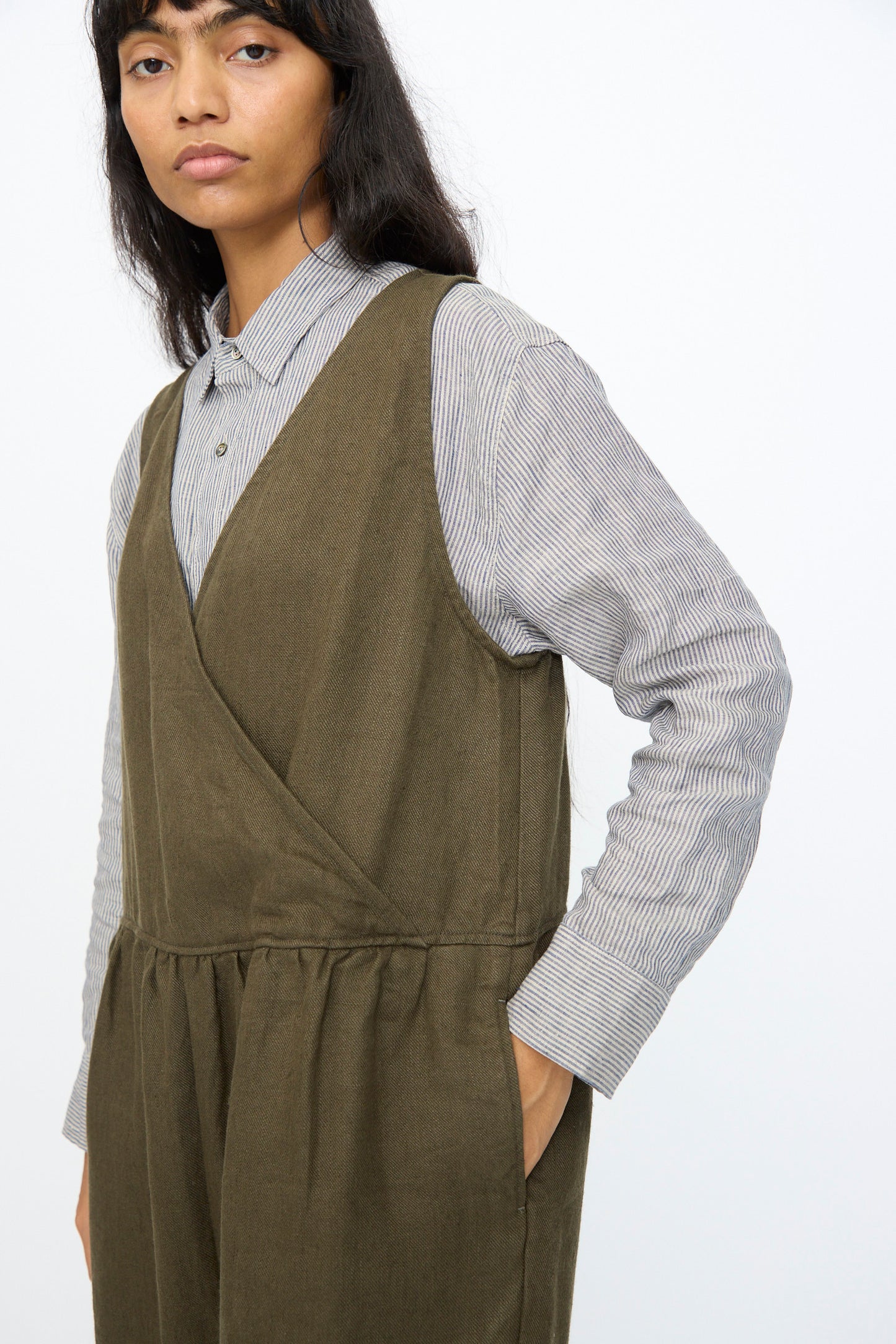 A person wearing the Ichi Antiquités Twill Weave Linen Jumpsuit in Khaki stands against a plain light background, layered over a gray striped button-up shirt.