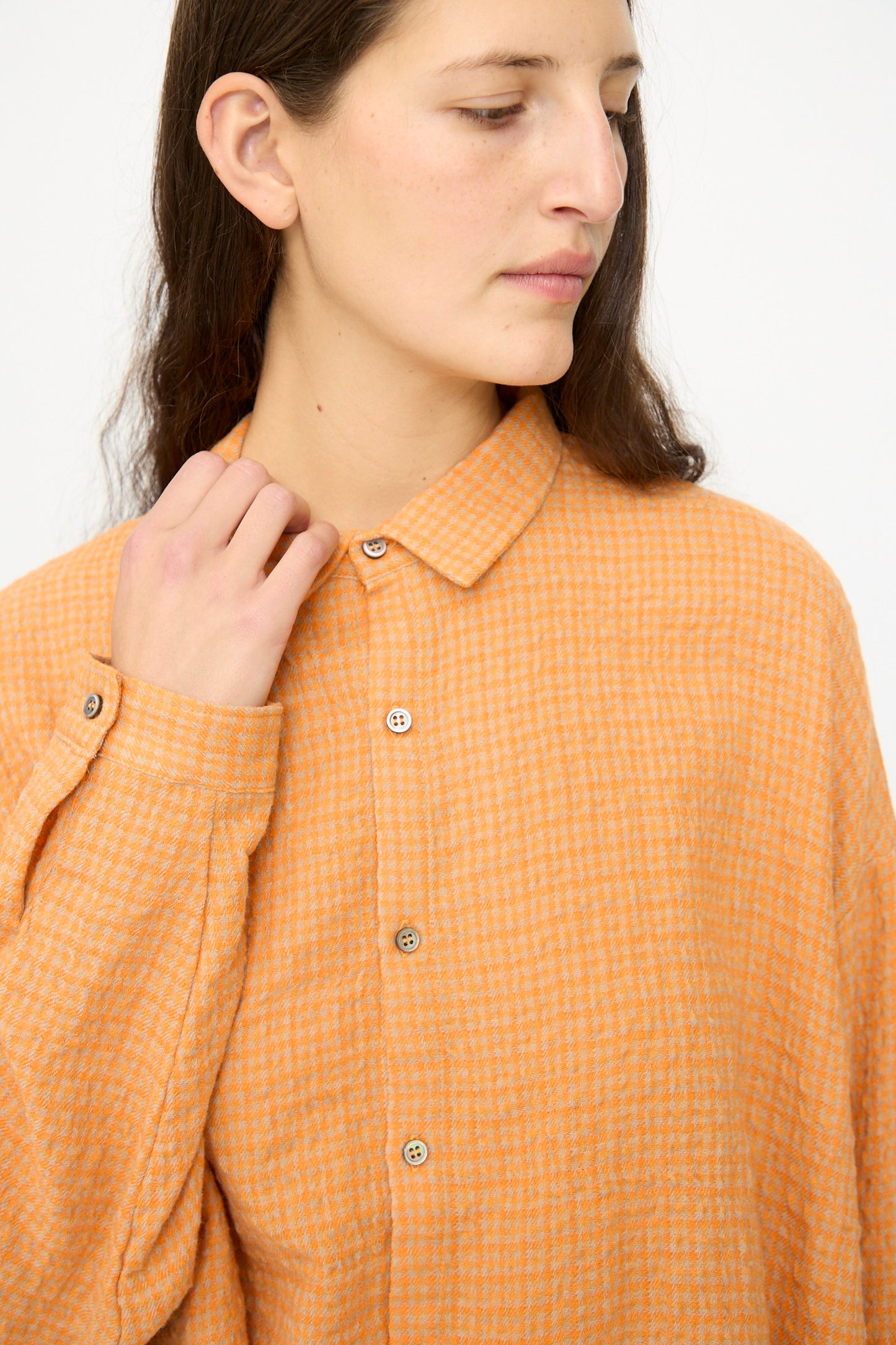 A person with long brown hair wears the Ichi Antiquités Wool Cotton Gingham Shirt in Orange and Mocha, made in Japan, looking down and to the side while holding the collar with one hand.