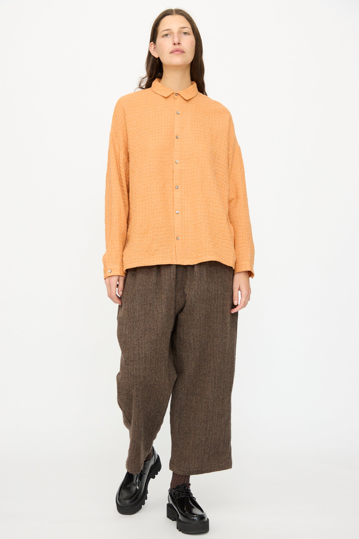 A person stands against a plain background wearing the Ichi Antiquités Wool Cotton Gingham Shirt in Orange and Mocha, paired with loose brown pants and black platform shoes.