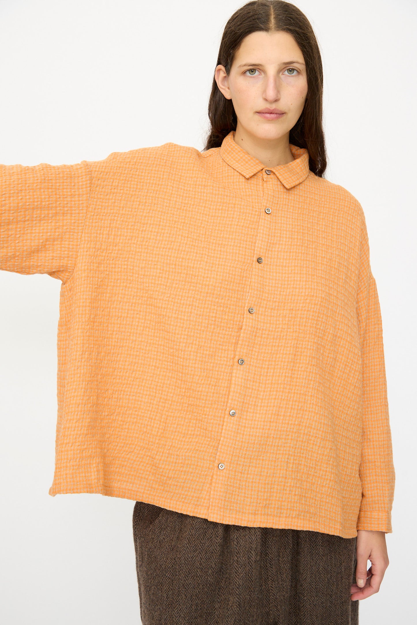 A person wears the Wool Cotton Gingham Shirt in Orange and Mocha by Ichi Antiquités, featuring long sleeves and an oversized fit, paired with brown pants. Made in Japan, they stand against a plain white background.