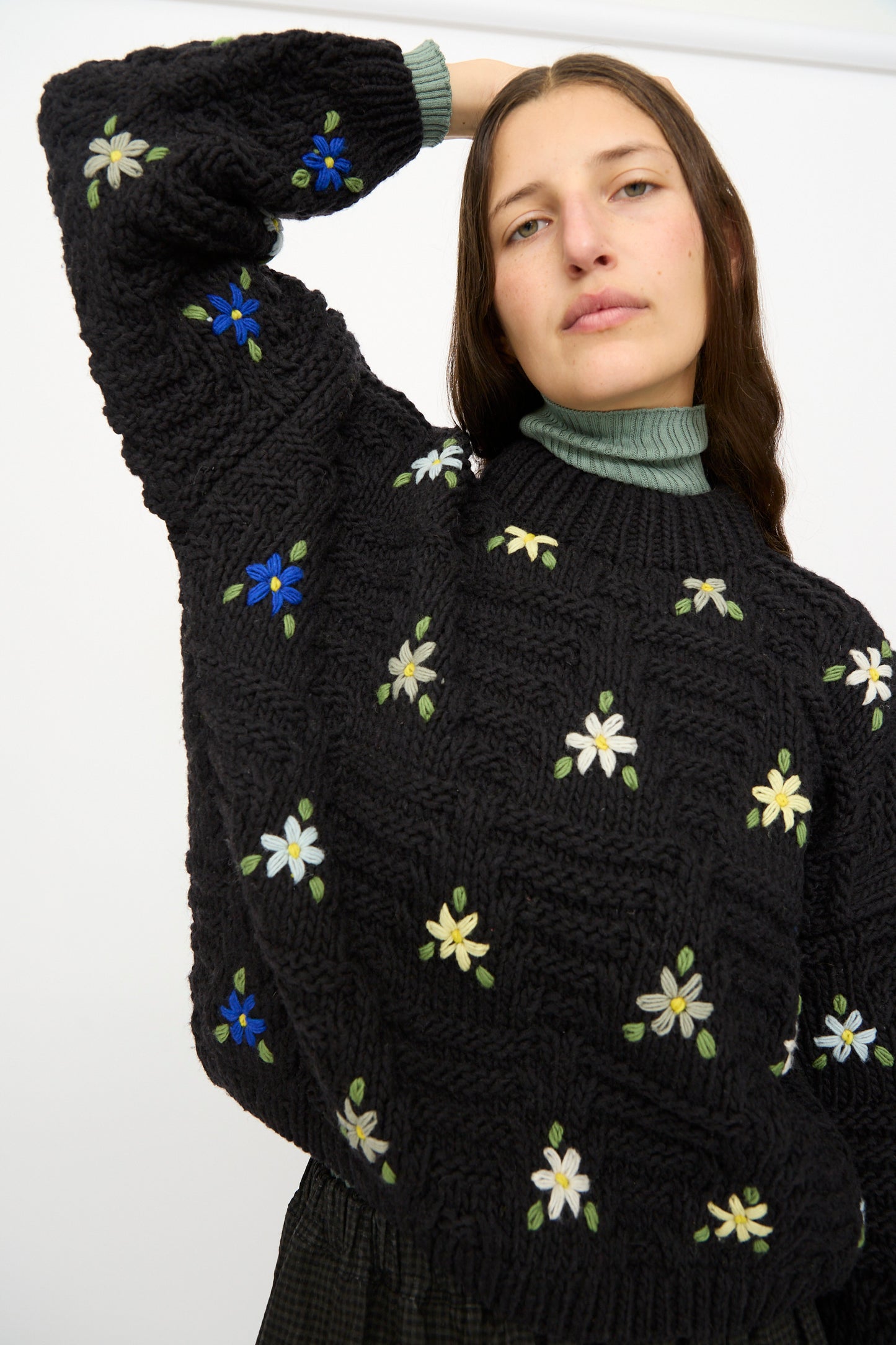 A person wears the Ichi Antiquités Wool Hand-knit Pullover with Flowers in Black, featuring colorful embroidered flowers, with one arm raised behind their head.