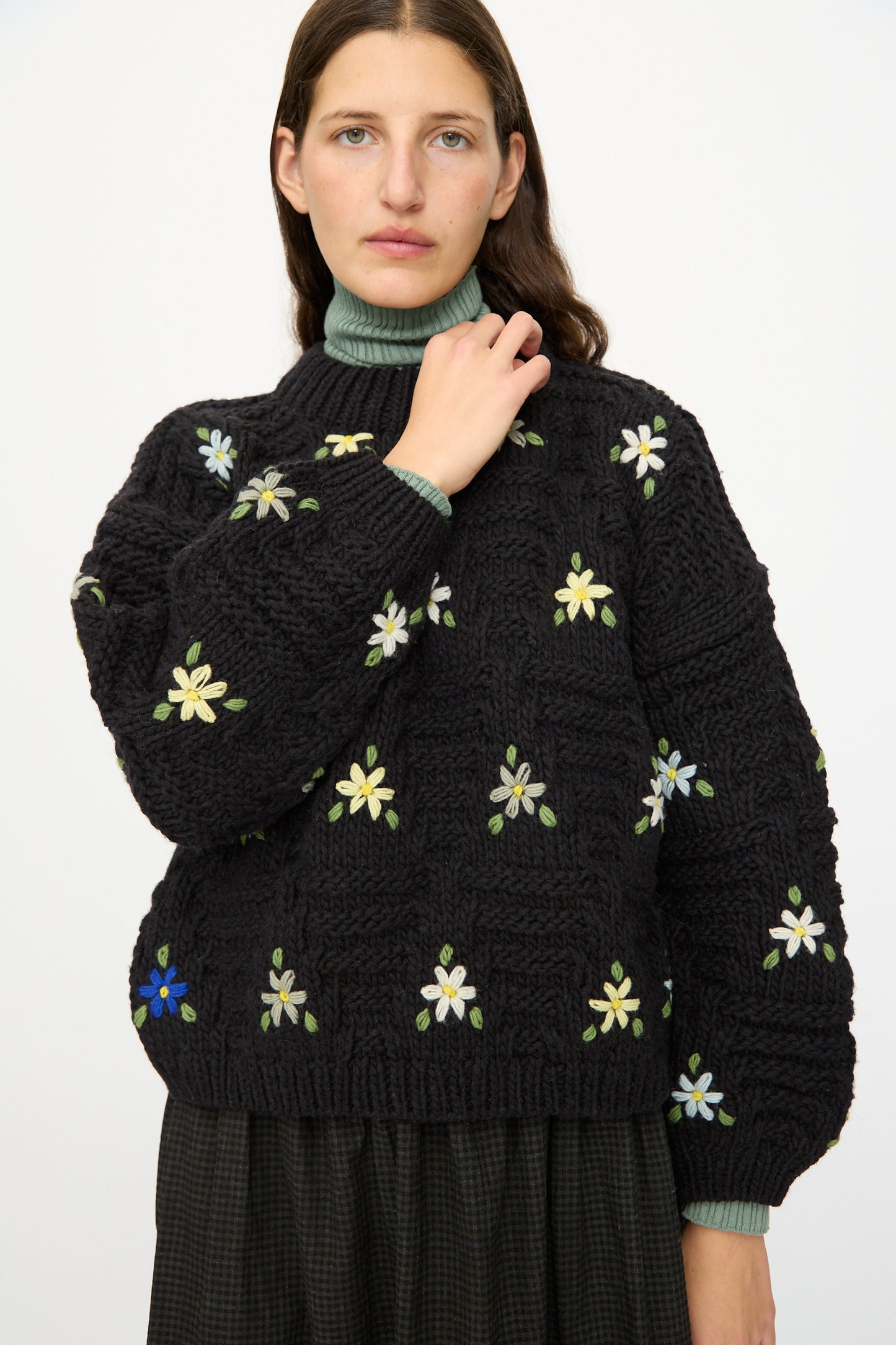 A woman with long brown hair models the Ichi Antiquités Wool Hand-knit Pullover with Flowers in Black, paired with a green turtleneck and dark skirt, standing against a plain white background.