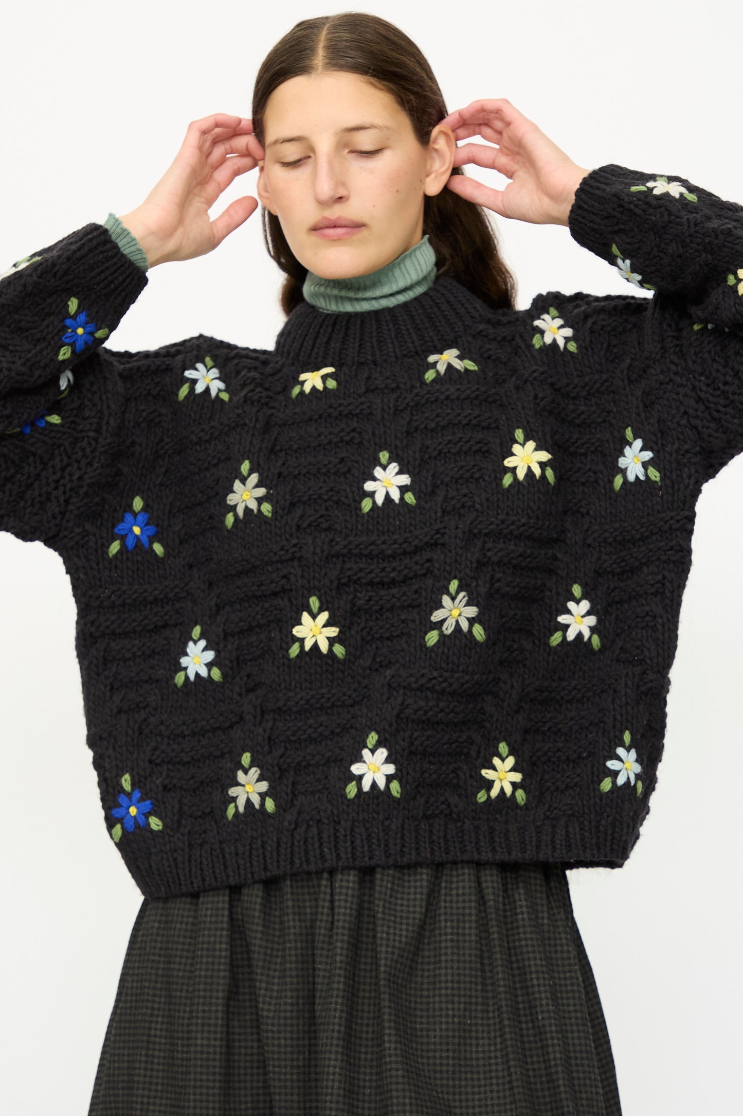 A woman stands with eyes closed, wearing the Ichi Antiquités Wool Hand-knit Pullover with Flowers in Black over a green turtleneck and a dark checkered skirt, her hands raised near her head.